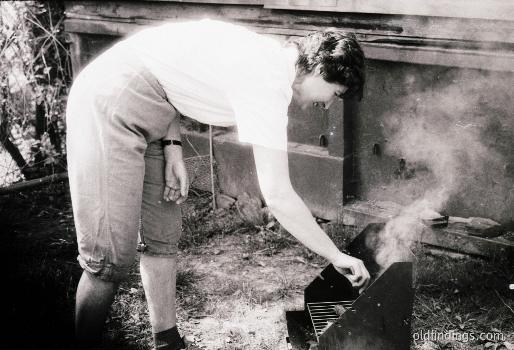 Black-and-white photo of two individuals tending a brick oven, likely for baking bread. The person on the right stirs embers with a metal tool, while the other stands nearby in rolled-up sleeves. Outdoor setting with visible smoke and rustic brickwork. Evokes mid-20th century domestic life, possibly rural Europe.