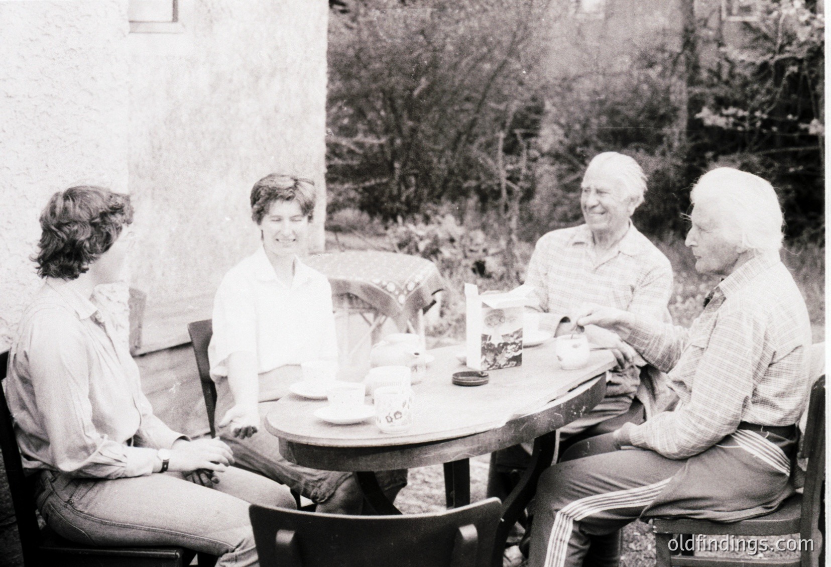 Four adults seated at an outdoor café table, mid-1970s. Lighting suggests daytime, with natural light filtering through trees. Men’s short-sleeved button-ups and women’s blouses reflect casual mid-century fashion. Ashtray and empty cups indicate a relaxed social gathering. é