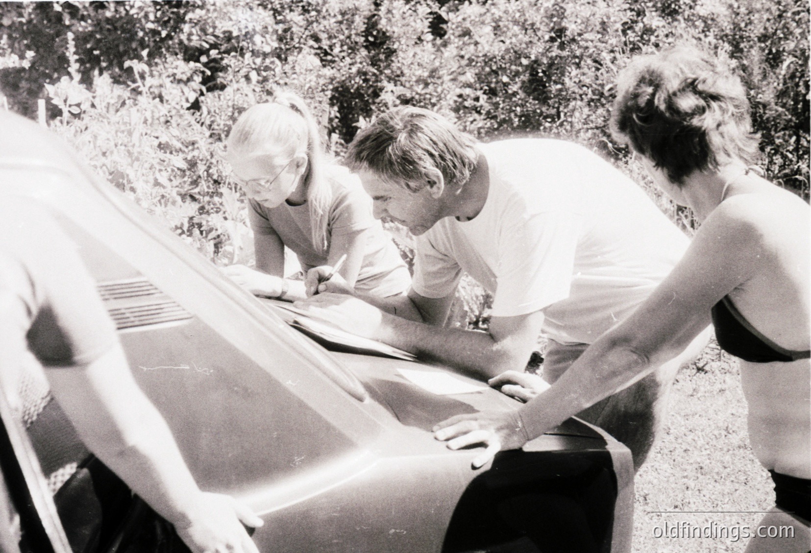 Three individuals engage in outdoor maintenance on a vintage sailboat, likely mid-20th century. The man in the center adjusts rigging while the woman on the left holds a line, and the woman on the right assists with a tool. Lush greenery and a wooden dock frame the scene, suggesting a coastal or lakeside setting.