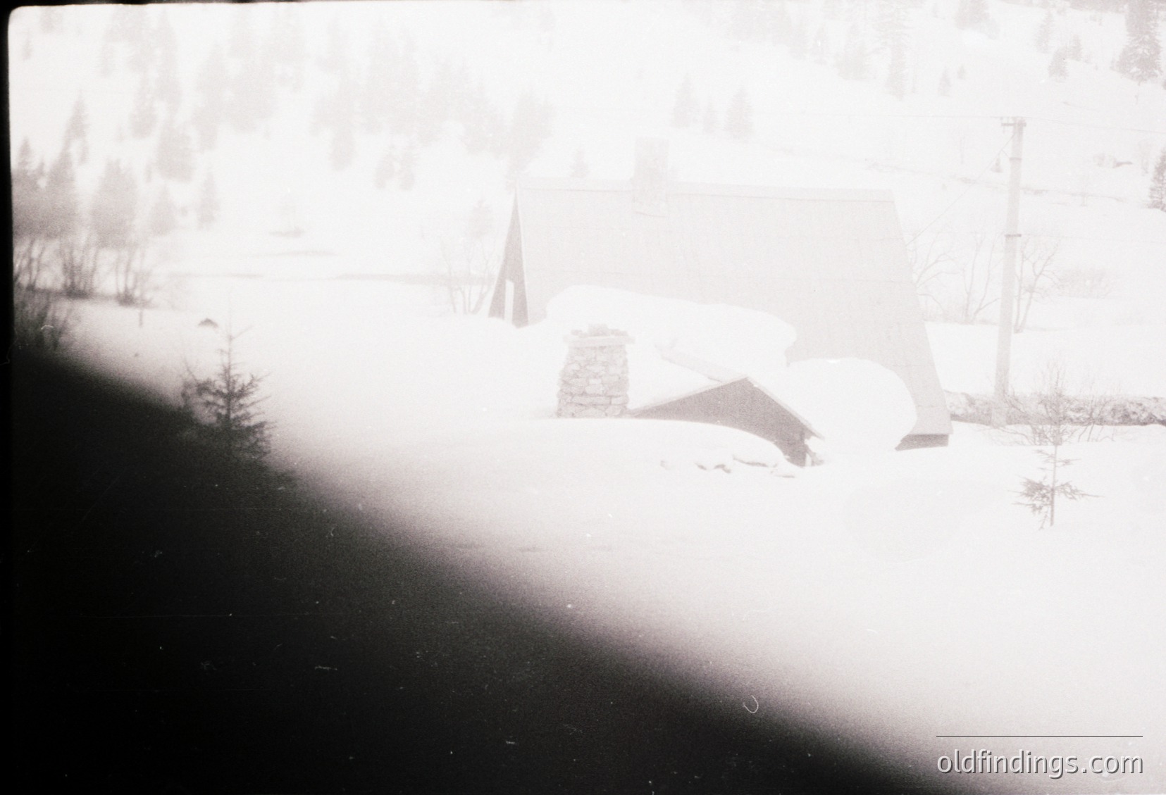 Vintage black-and-white photo of a rural landscape with snow-covered ground and sparse trees. A lone, partially buried structure—likely a wooden shed or outbuilding—sits near a flagpole. Overcast sky and utility poles suggest mid-20th century rural Europe.