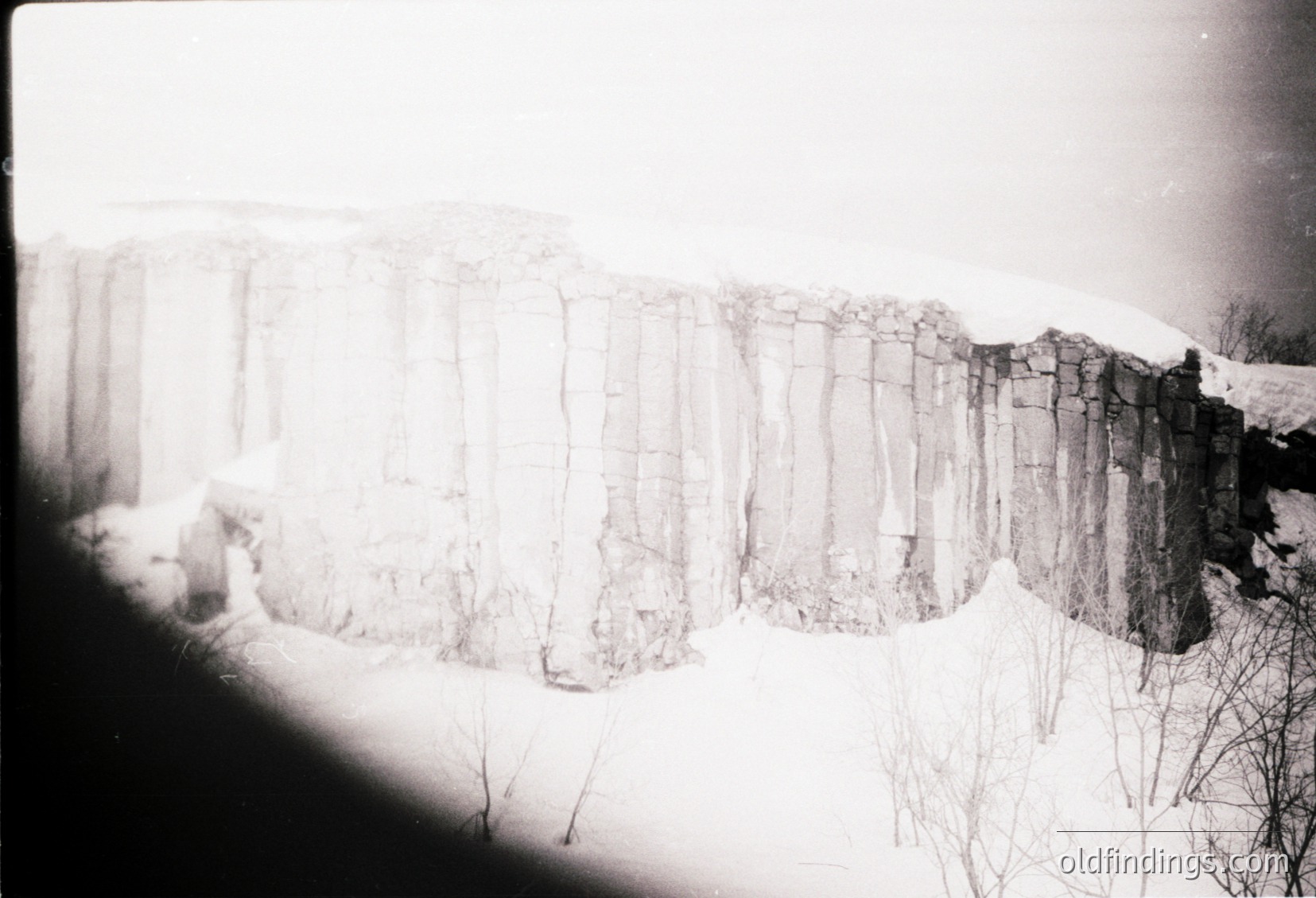 Vintage black-and-white shot of a stone wall with vertical, evenly spaced bricks—likely a historic or agricultural boundary. Snow covers the ground and wall, suggesting winter. Minimalist composition with stark contrast.
