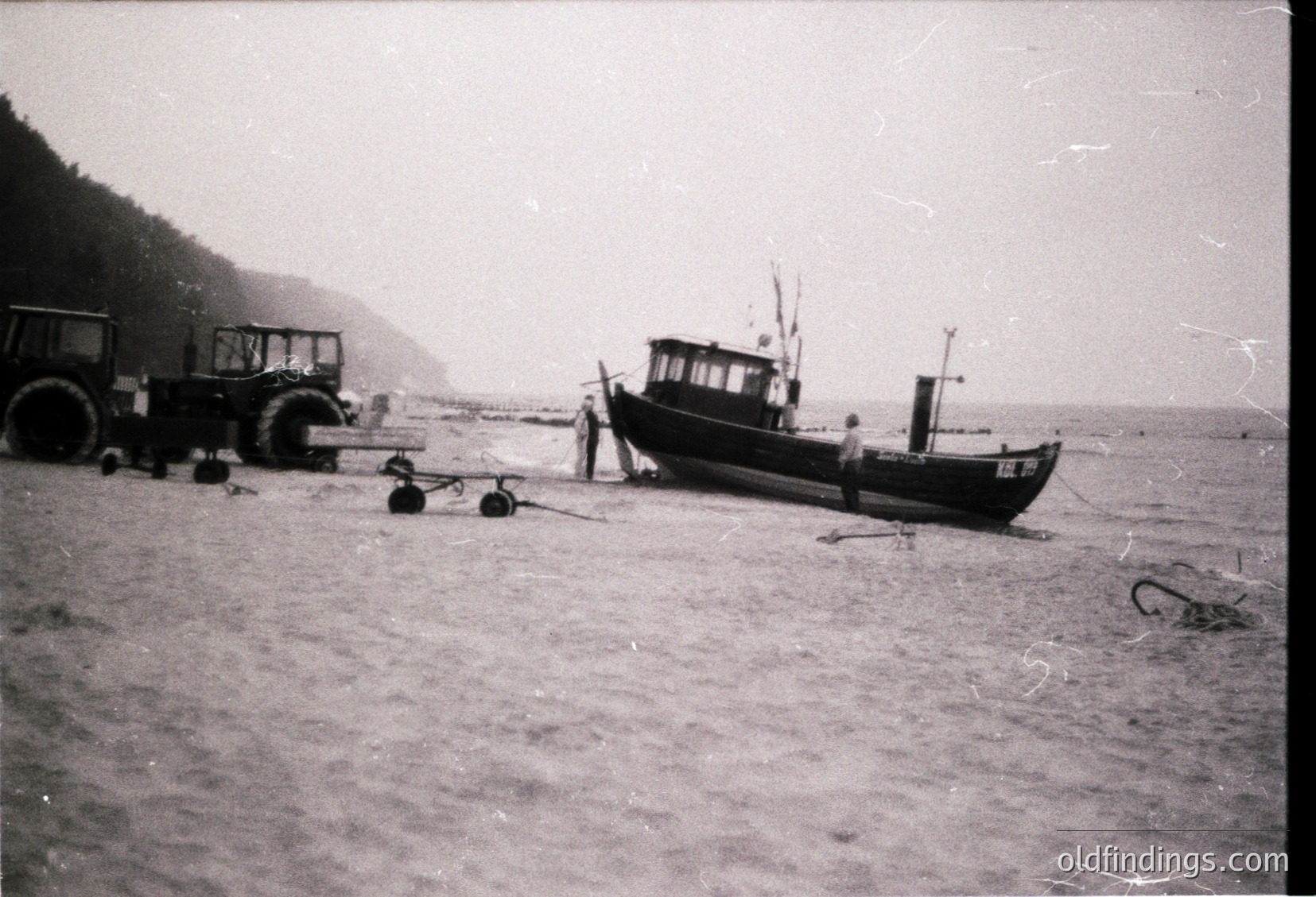 Vintage black-and-white coastal scene featuring a small wooden fishing boat grounded on a sandy shore. Two tractors (likely agricultural) positioned near the water’s edge, suggesting port or dockside activity. A lone figure inspects the boat’s hull. Rugged cliffs and misty horizon hint at a remote seaside location. Mid-20th century industrial maritime heritage.