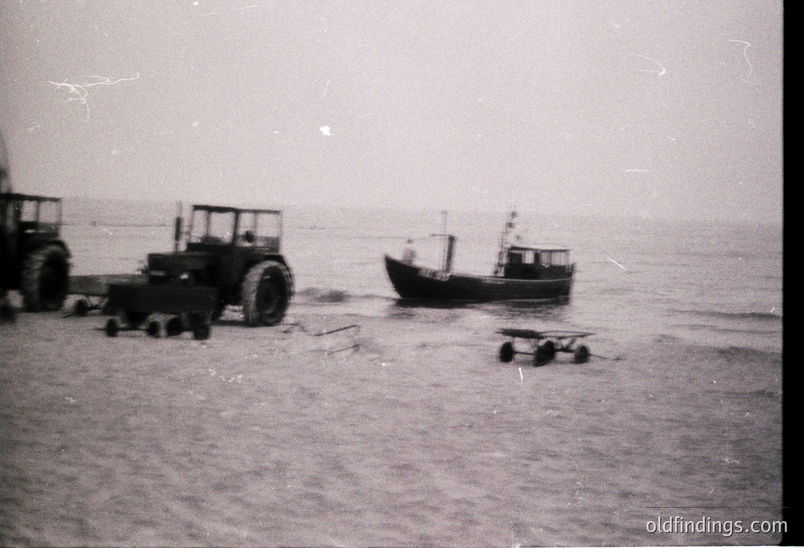 Vintage black-and-white coastal scene featuring a small fishing boat grounded on sandy shore, flanked by industrial tractors and wooden pallets. Likely mid-20th century industrial port activity.