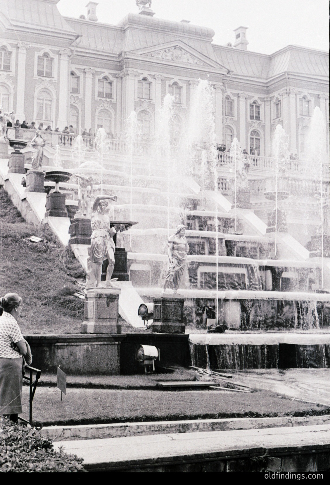 Baroque-style fountain with cascading waterfalls and sculpted figures, set against grand neoclassical architecture. Mid-20th century (likely 1950s–60s) urban park scene. Visitors in casual attire—one seated, others observing or walking near the water.