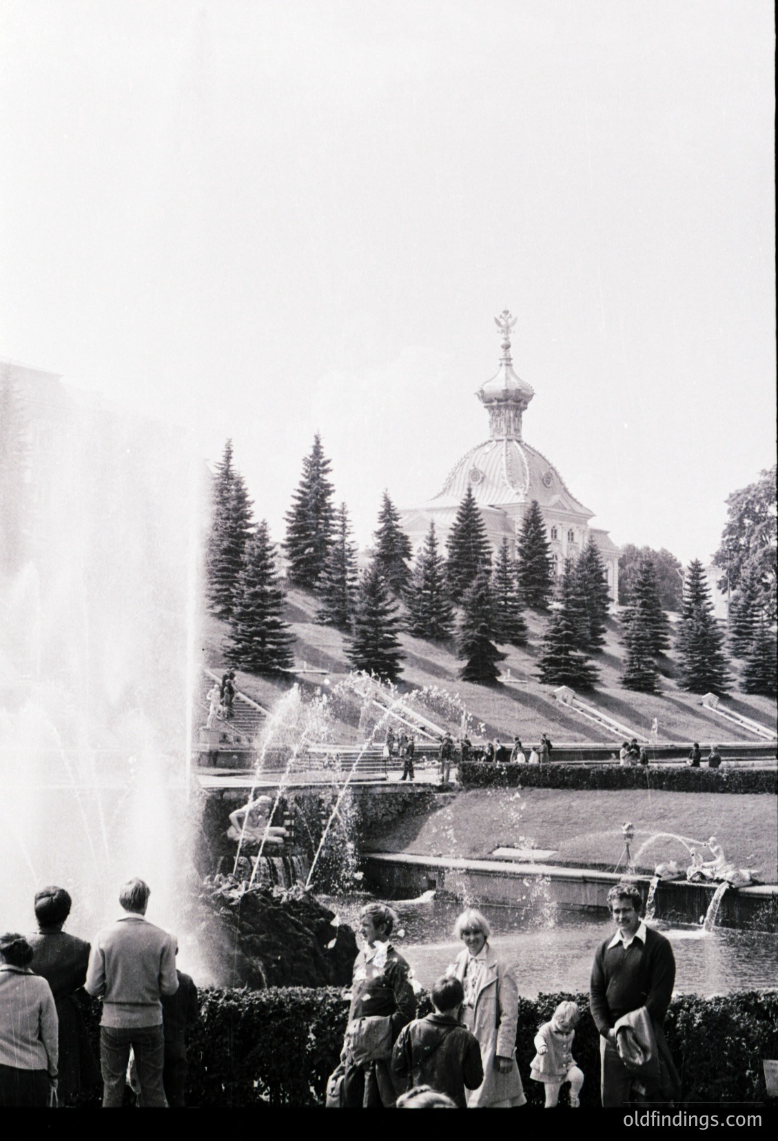 Vintage black-and-white shot of a grand fountain in a landscaped park, with a prominent Orthodox church featuring onion domes in background. Mid-20th century attire suggests 1950s–1960s era. Crowd includes adults and children observing water jets.