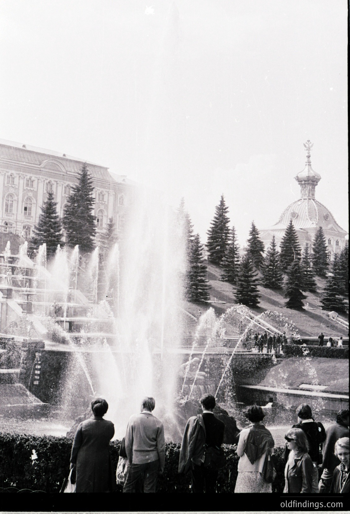 Black-and-white shot of a grand fountain in a public park, likely Soviet-era architecture. Multi-tiered water jets cascade toward a central spout, framed by coniferous trees and ornate buildings. Crowd in 1960s-70s attire—men in suits, women in dresses—watches.