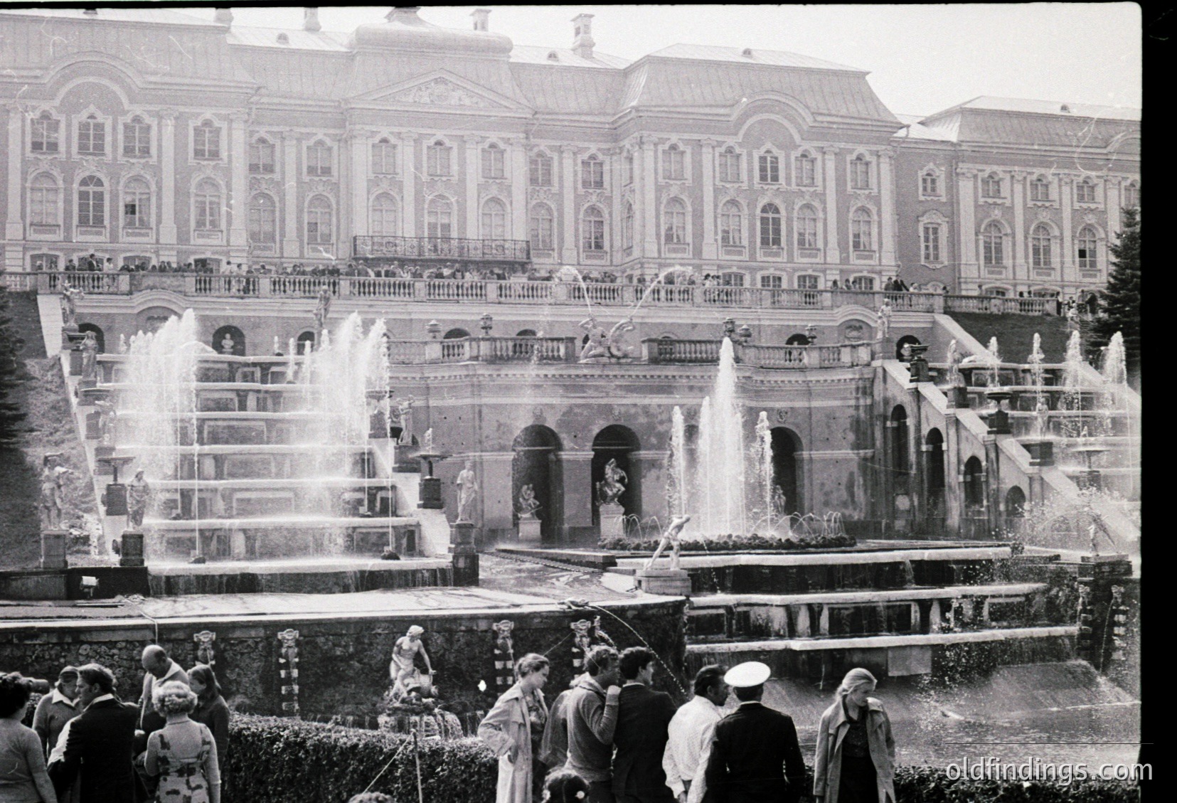 Baroque-style palace courtyard with cascading tiered fountains and ornate statues, likely European. Mid-20th century attire suggests 1950s–1960s era. Crowds gather around water features, indicating public or tourist access.