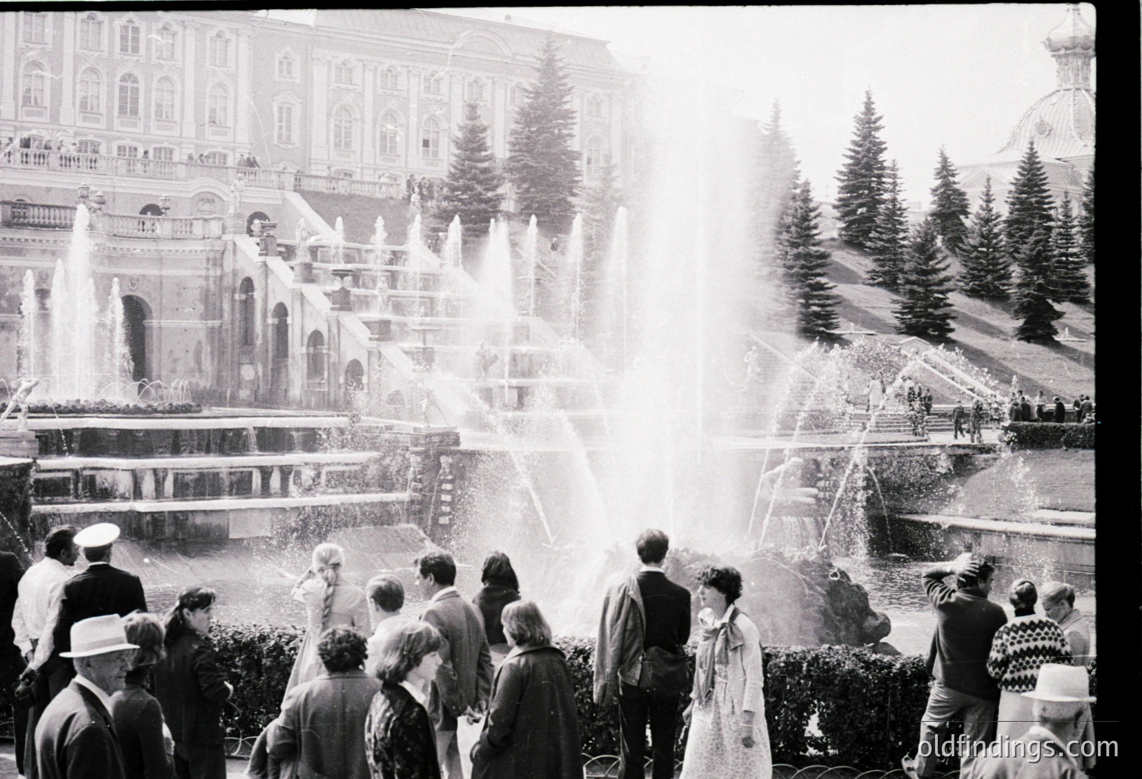 Grand tiered fountain in a grand alpine resort, surrounded by elegant 1950s–60s architecture. Crowds in winter attire—hats, coats—gather around the cascading water. Snow-capped pines frame the scene, hinting at a mountainous European location.