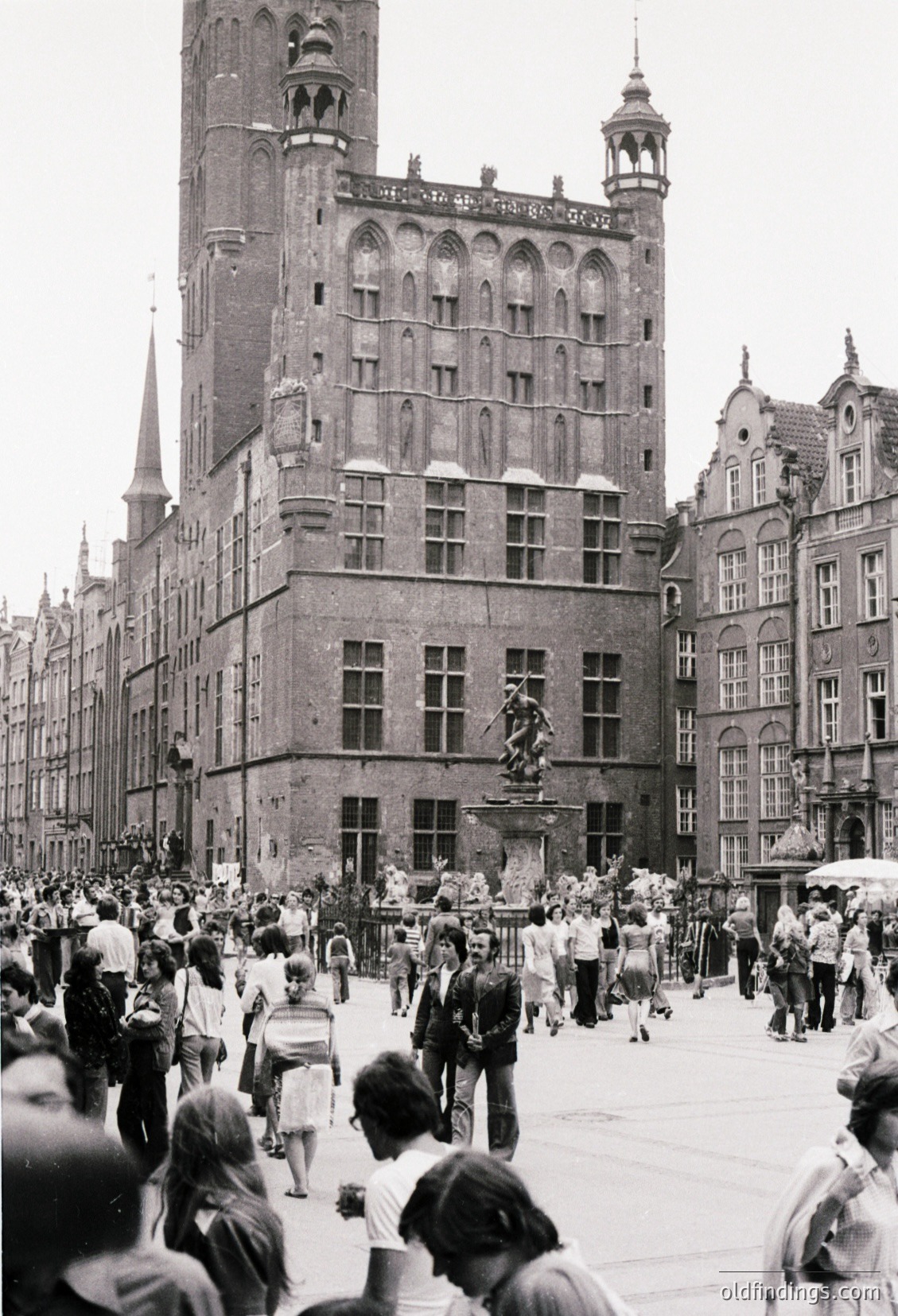Historic European square bustling with mid-20th century attire. Gothic-style brick buildings with ornate facades and spires dominate the scene. Central statue on pedestal likely commemorates a notable figure. Crowd of casually dressed pedestrians, suggesting a public gathering or tourist area.