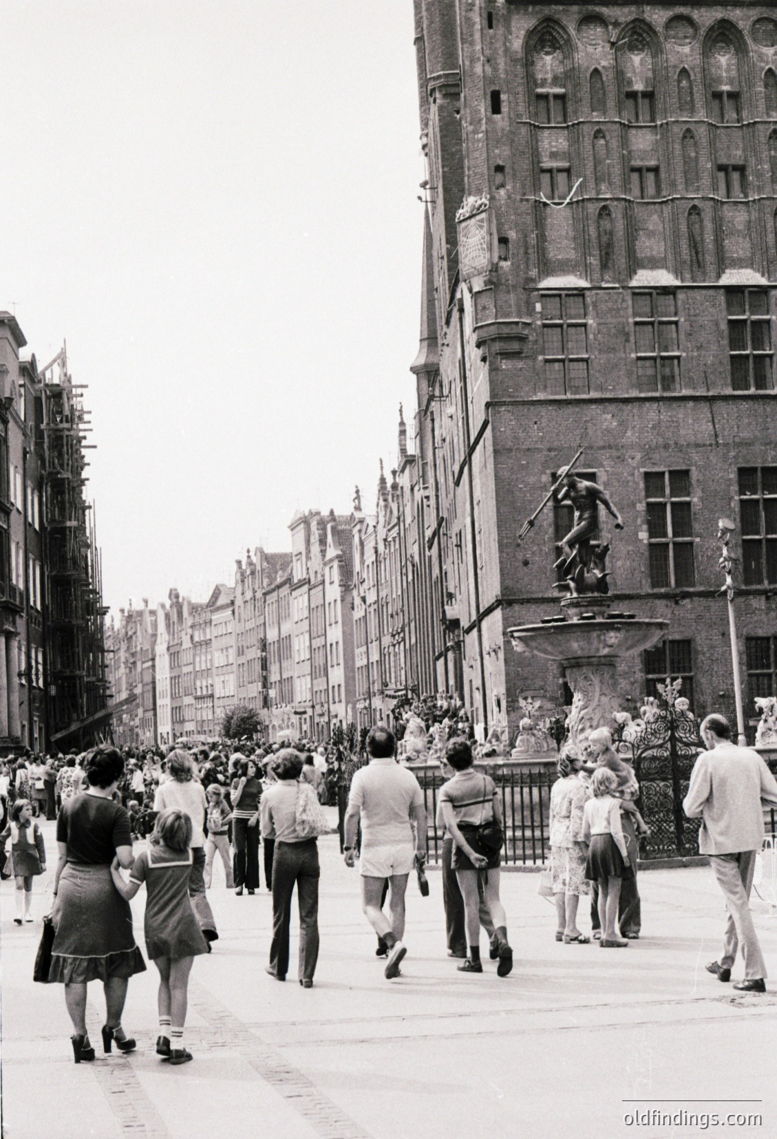Vintage street scene featuring Gothic Revival architecture with ornate stonework and a prominent fountain statue. Crowd of casually dressed pedestrians, likely 1950s–1960s, walking along a wide, open plaza. Mid-century fashion includes knee-length skirts, short-sleeve shirts, and flat shoes. Urban European setting with historic buildings lining both sides.