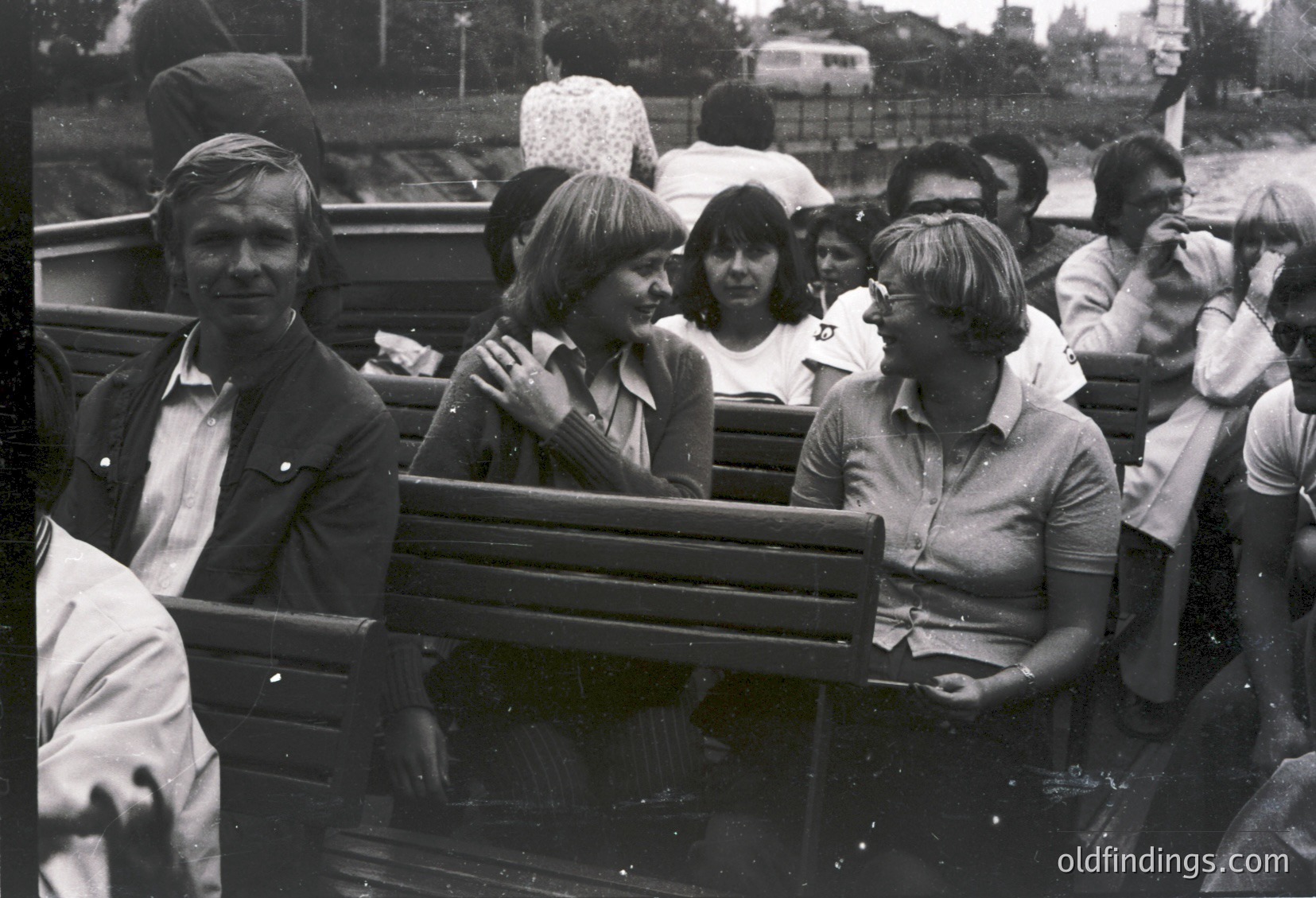 Black-and-white photo of a group seated in an open-air tram or trolley, likely from the **1960s-1970s**. Urban setting with blurred buildings and greenery in background. Men and women in casual 1970s attire—short-sleeved shirts, headscarves, and light jackets. One man wears a cap; another holds a camera. Crowded, informal atmosphere suggests a public outing or tourist activity.