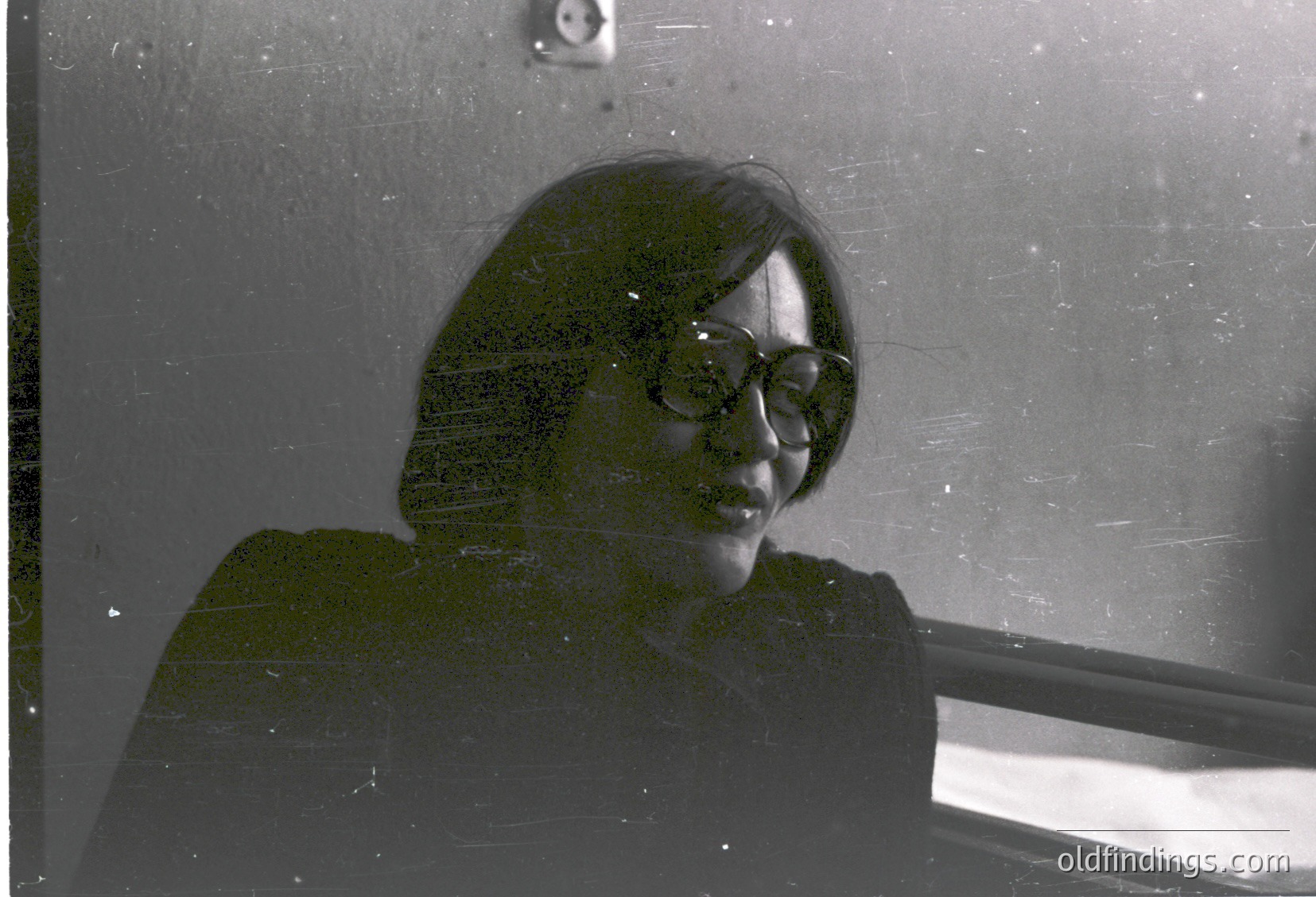 Vintage black-and-white portrait of a person viewed through a fogged or scratched window, evoking mid-20th-century documentary style. Short hair, round glasses, and a neutral expression. Indoor setting with muted lighting, suggesting a candid or candid-style photograph.