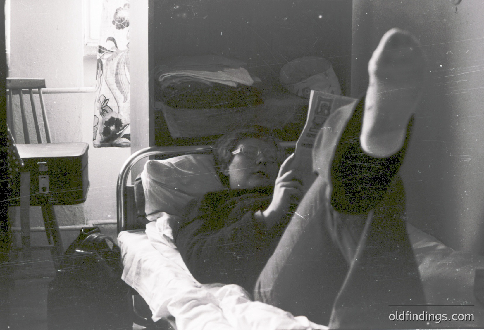 Vintage black-and-white photo of elderly person reclining in hospital bed, reading newspaper. Visible medical equipment (IV stand) and floral wall mural. Likely 1960s–1970s healthcare setting.