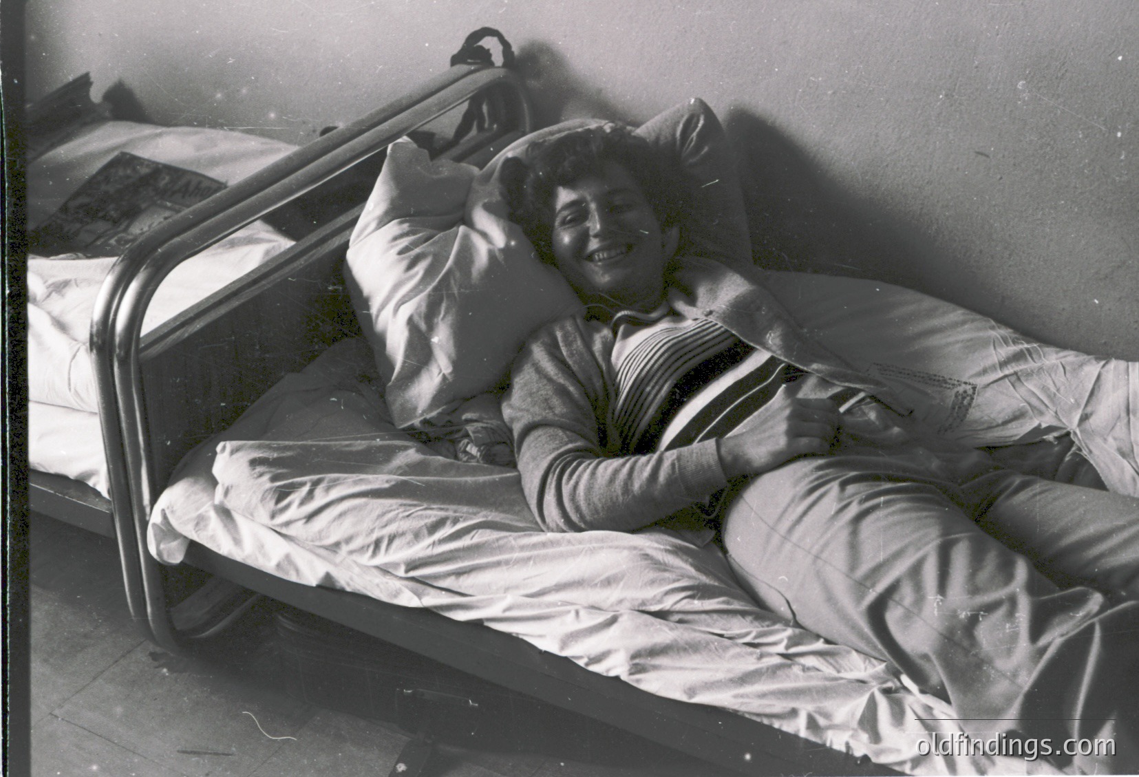 Vintage black-and-white hospital bed with metal railings, showing a relaxed individual in striped pajamas resting on pillows. Soft lighting suggests indoor recovery setting, likely mid-20th century.