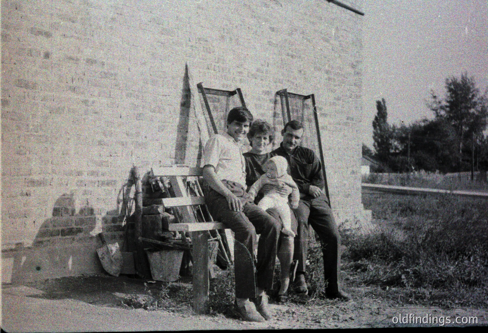 Three individuals pose outdoors on a weathered concrete bench beside a brick wall, likely mid-20th century. The man on the left wears a short-sleeve shirt and dark trousers; the woman in the center holds a child in a light dress; the man on the right wears a tie and dark jacket. A fishing net leans against the wall, suggesting recreational activity. Overgrown vegetation and a blurred background hint at a rural or semi-rural setting.