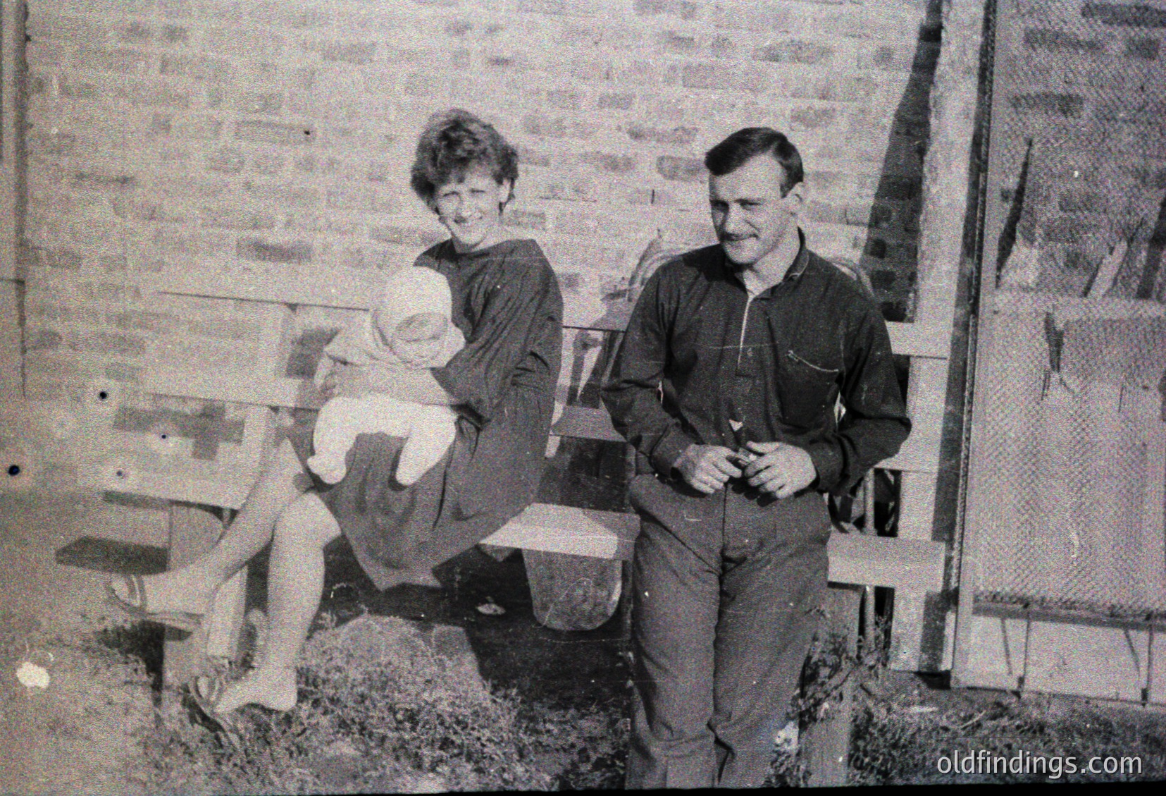 Family portrait featuring a woman holding a baby on a rustic wooden bench beside a brick wall, with a man standing beside her. Mid-20th century rural or suburban setting, likely 1950s–1960s.