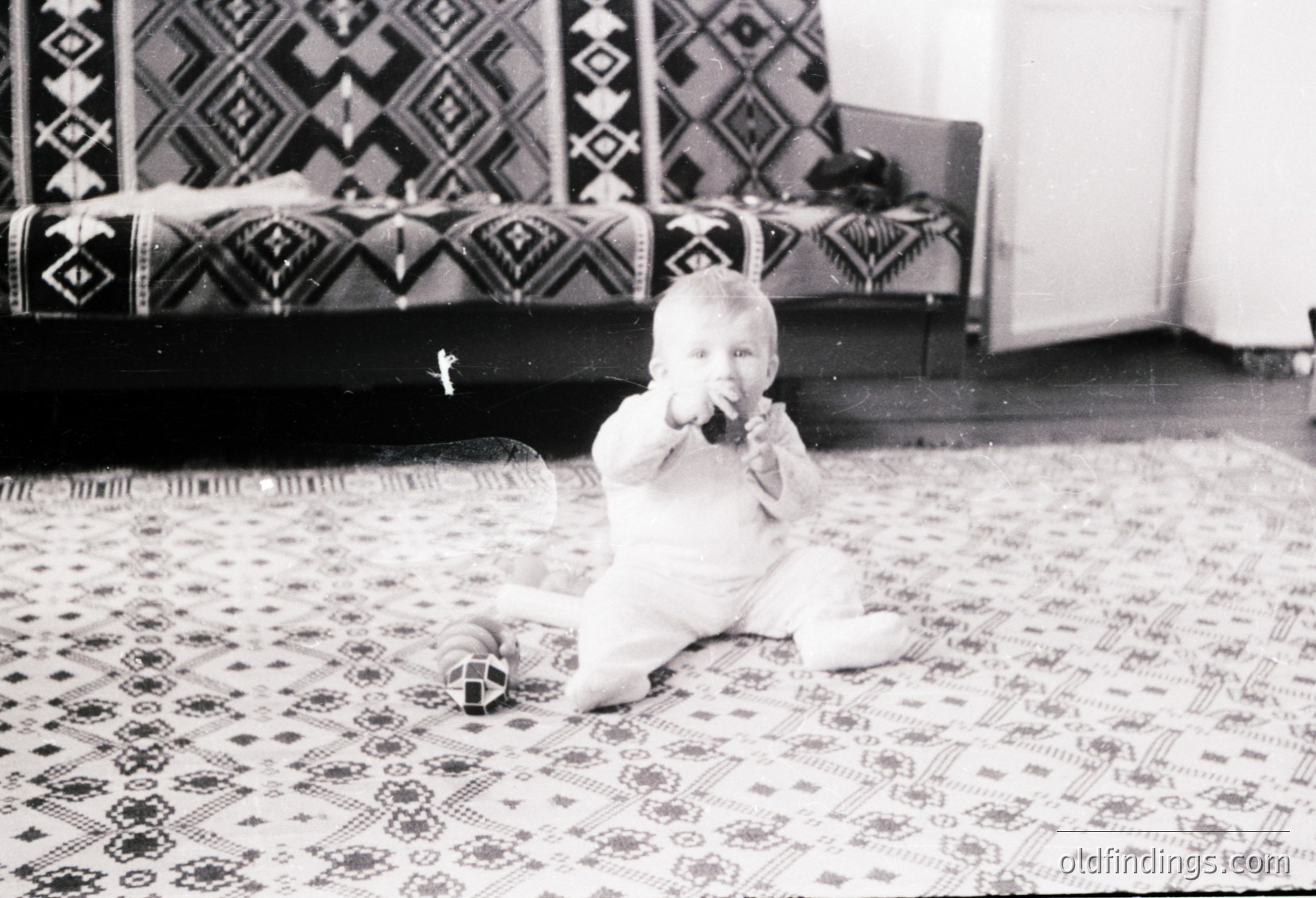 Vintage black-and-white photo of an infant playing on an intricately patterned rug, holding a toy. Geometric textile designs on sofa and floor suggest mid-20th century home decor. Soft focus and candid composition evoke nostalgia.