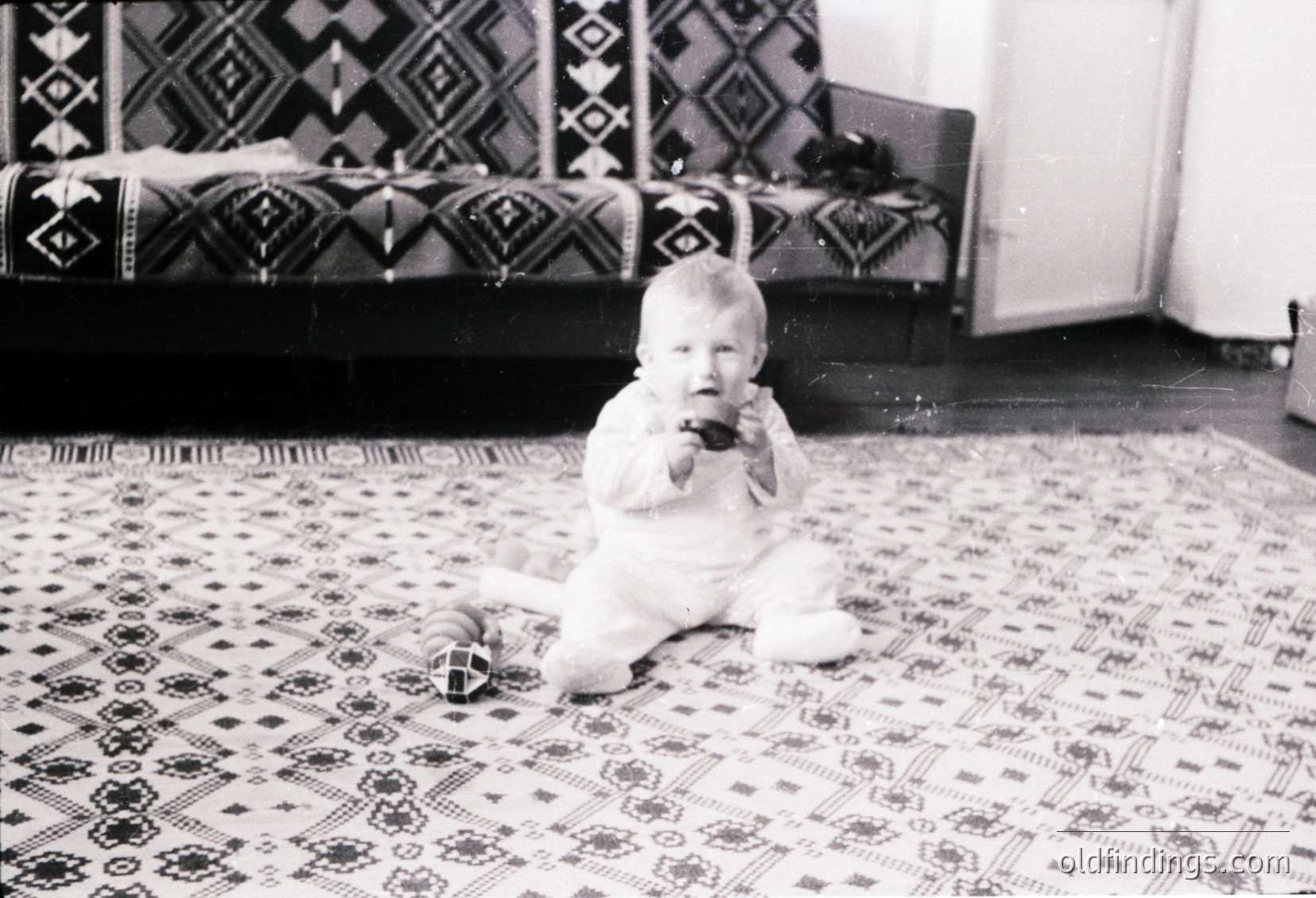 Black-and-white snapshot of an infant in a white knitted outfit, sitting on an intricately patterned rug with geometric designs. The setting appears domestic, likely mid-20th century. Traditional textiles and simple furniture suggest a modest, culturally rich interior.