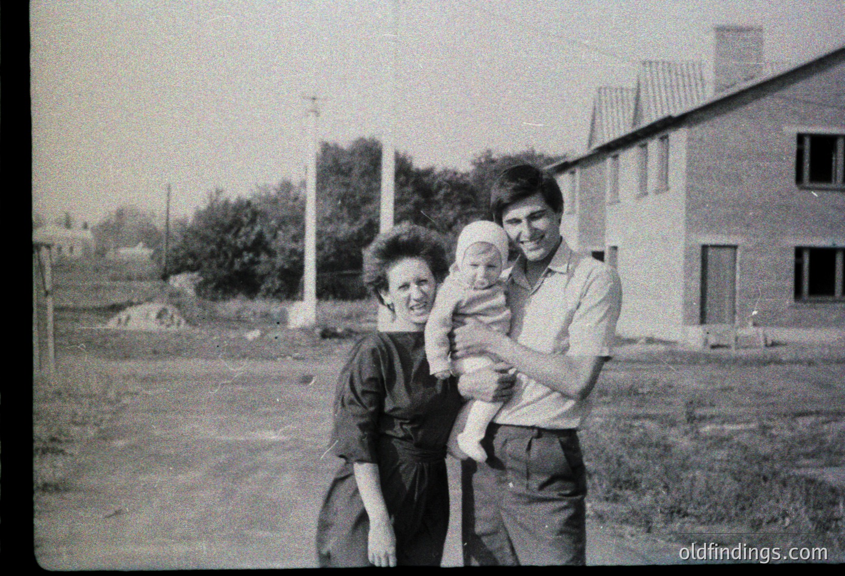 A mid-20th-century black-and-white family portrait: a woman cradles a baby, a man stands beside her in a grassy residential area with unfinished houses in the background.