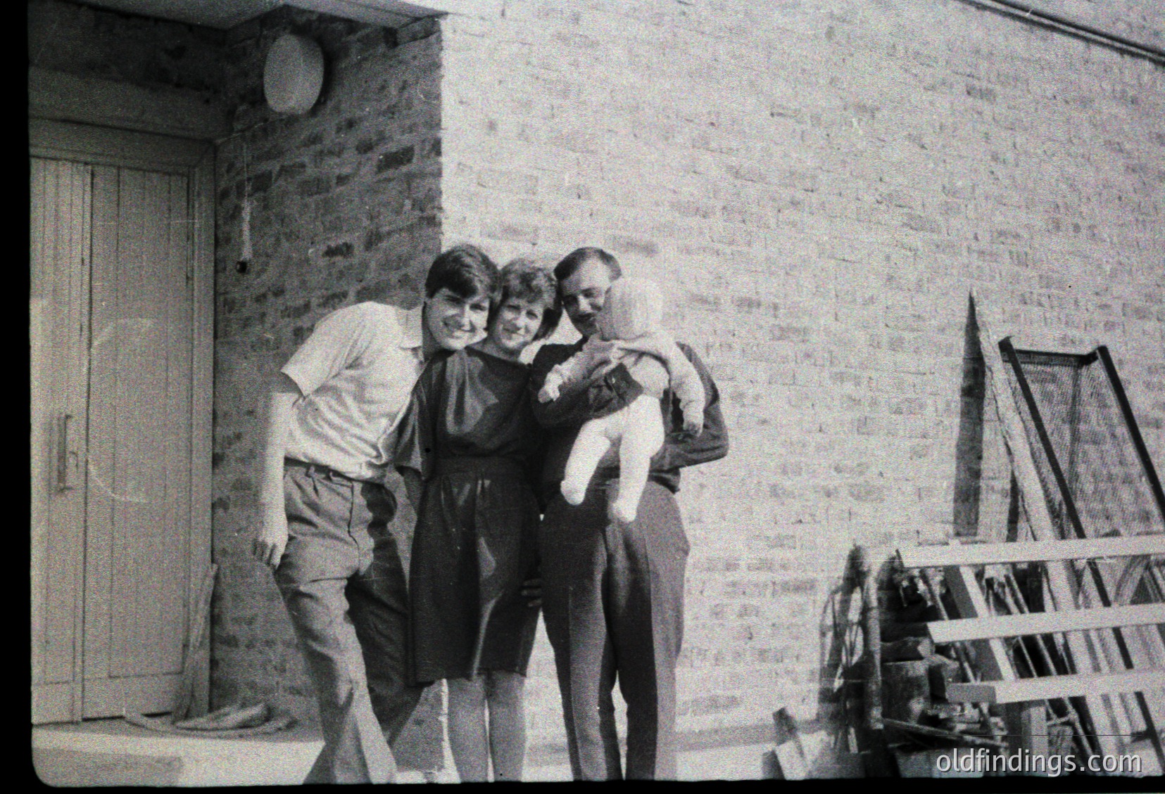 Family portrait outside a brick building, likely residential, with a wooden door and ladder leaning against the wall. Three adults and a baby in 1960s-1970s attire: man in sweater & trousers, two women in knee-length dresses, and a baby in a white onesie. Warm, candid moment capturing mid-century family life.