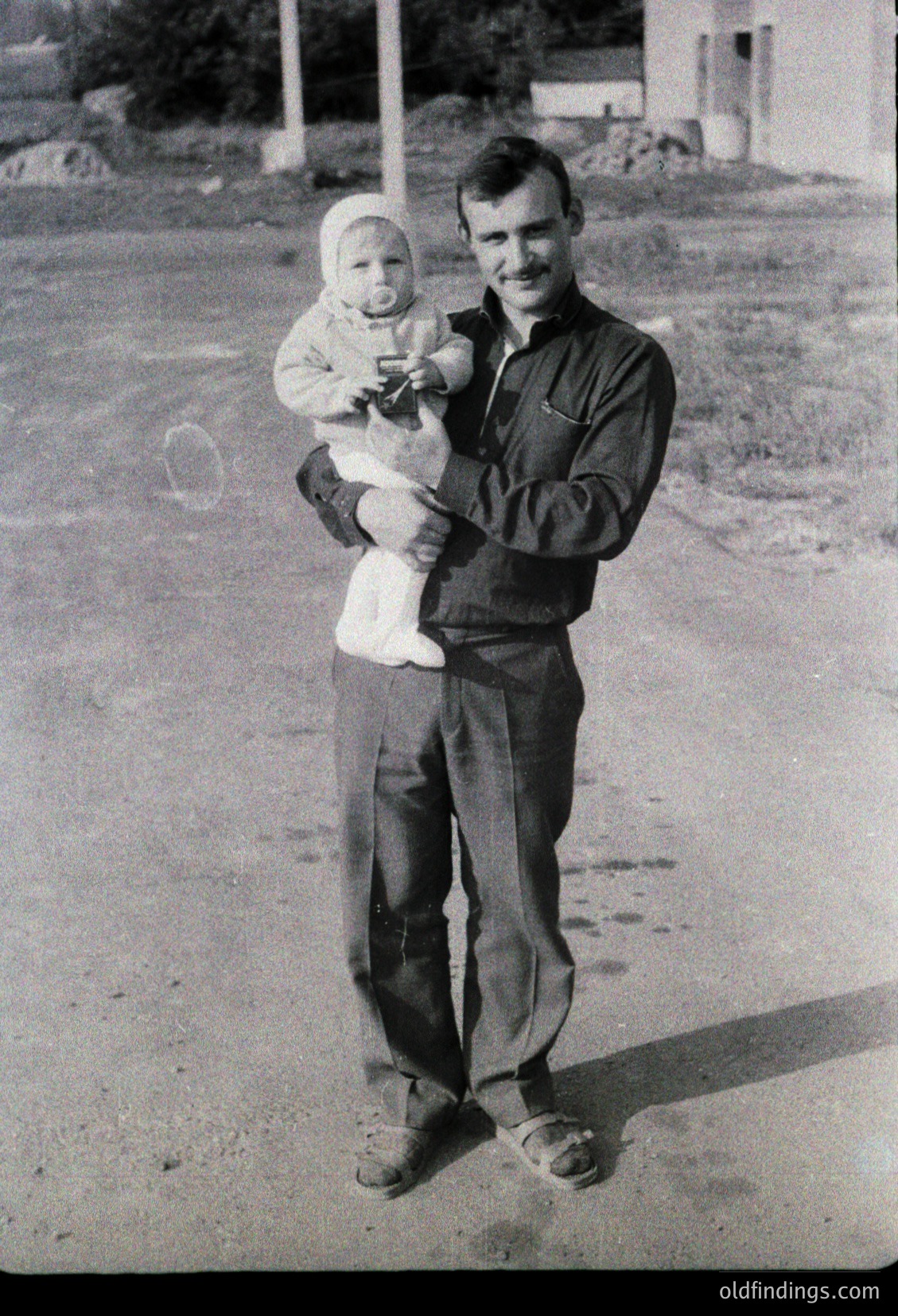 Mid-20th century black-and-white portrait of a man holding an infant outdoors, likely 1950s–1960s. Man wears button-up shirt, trousers, and loafers; infant in a white dress. Gravel road and simple residential background suggest rural or suburban setting. Candid, nostalgic family moment.