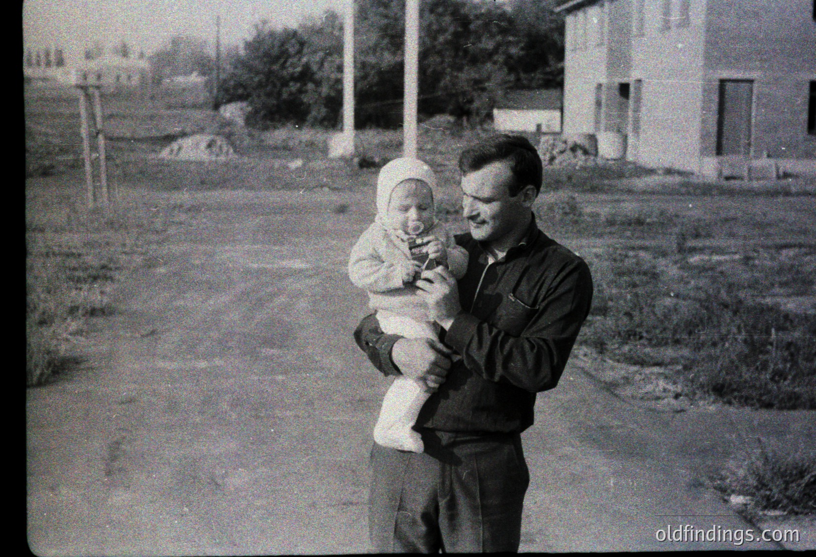 A man in mid-20th-century attire holds a baby outdoors in a residential area. The man wears a dark jacket and glasses; the baby is bundled in a white headscarf and light sweater. Suburban houses and overgrown grass frame the scene, suggesting a mid-century American or European setting.