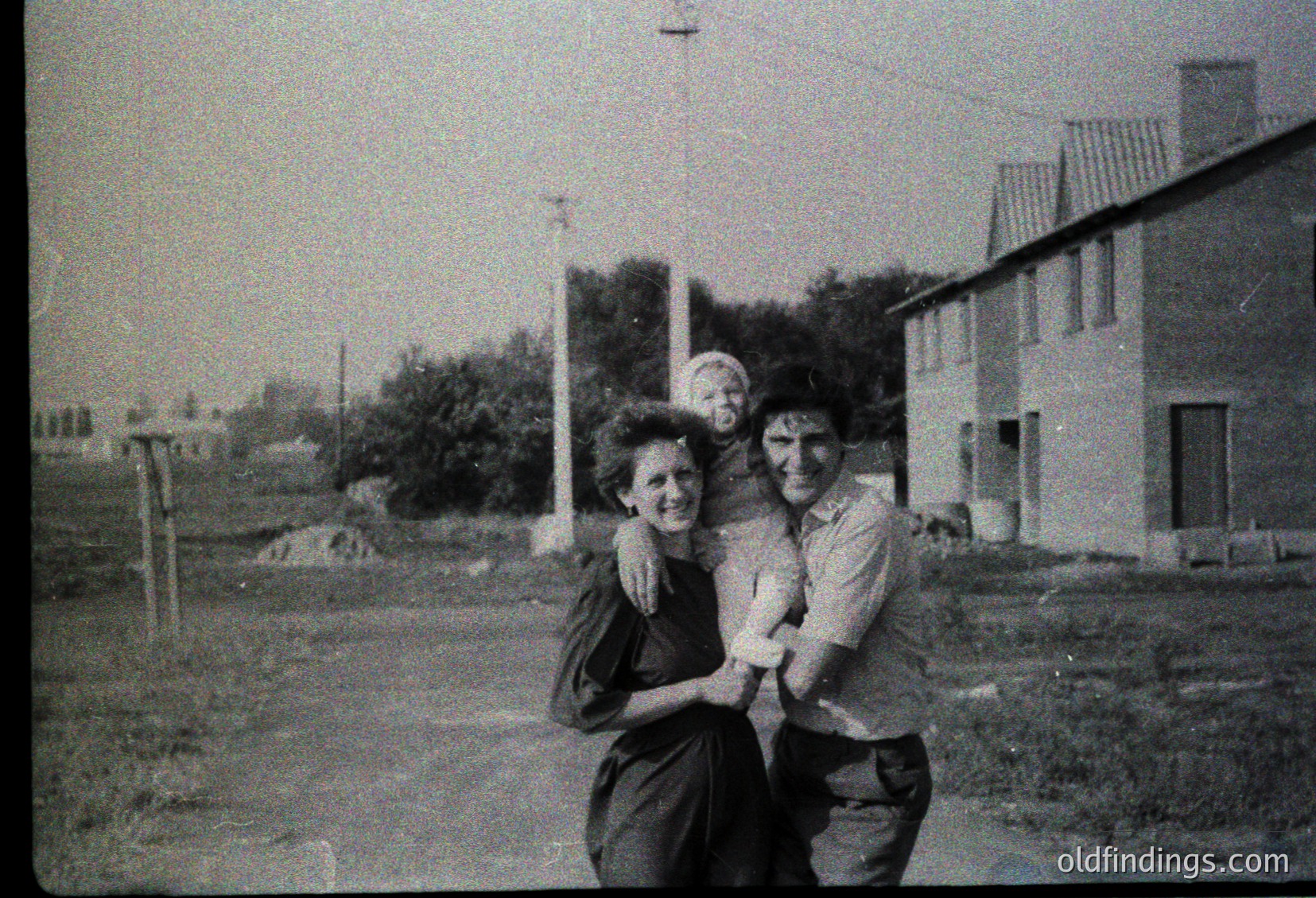 A black-and-white snapshot of three individuals posing outdoors in a rural setting, likely mid-20th century. The woman and man embrace, while a child stands between them. Simple residential buildings with flat roofs and a utility pole frame the scene. Overgrown vegetation and unpaved ground suggest a modest, possibly agricultural area.