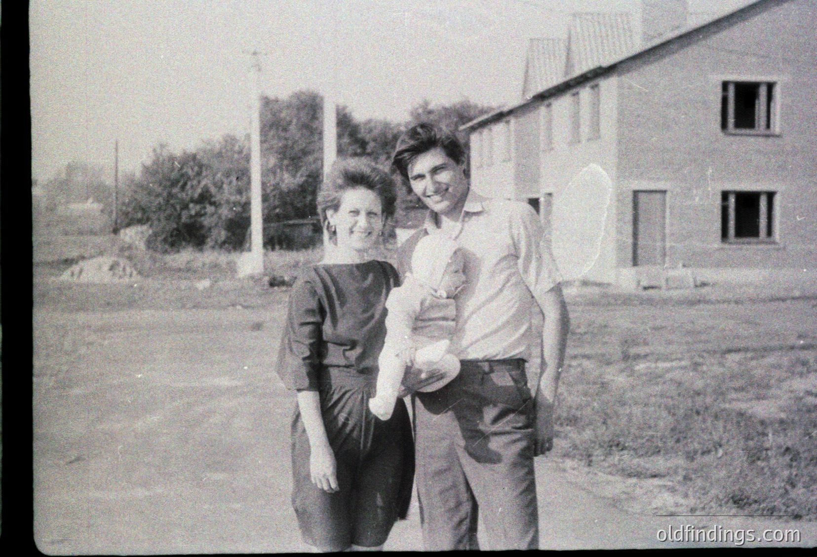Vintage black-and-white photo of two adults posing outdoors with a child, likely mid-20th century. Woman in fitted dress, man in button-up shirt and rolled pants holding a baby. Plain concrete buildings and open grassy area in background.