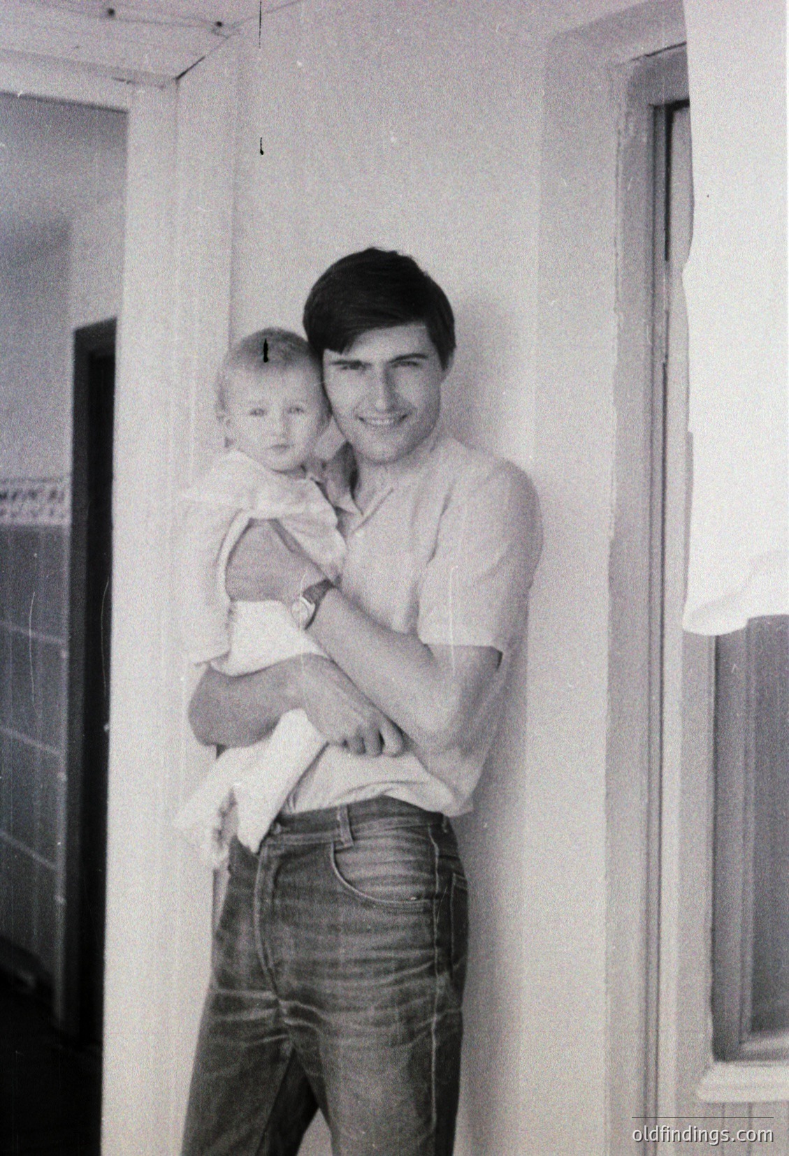 A man in a short-sleeved button-up shirt and jeans cradles a baby indoors, likely mid-20th century. Plain walls and a window frame suggest a modest residential setting. The composition captures a tender, candid family moment.