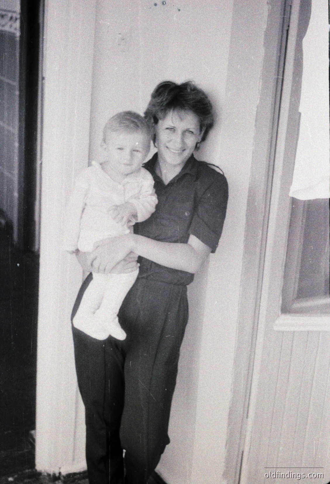 A woman in 1970s-era dress and trousers holds a toddler in a simple white shirt and diaper against a plain interior wall. Warm, candid moment captures generational bond.