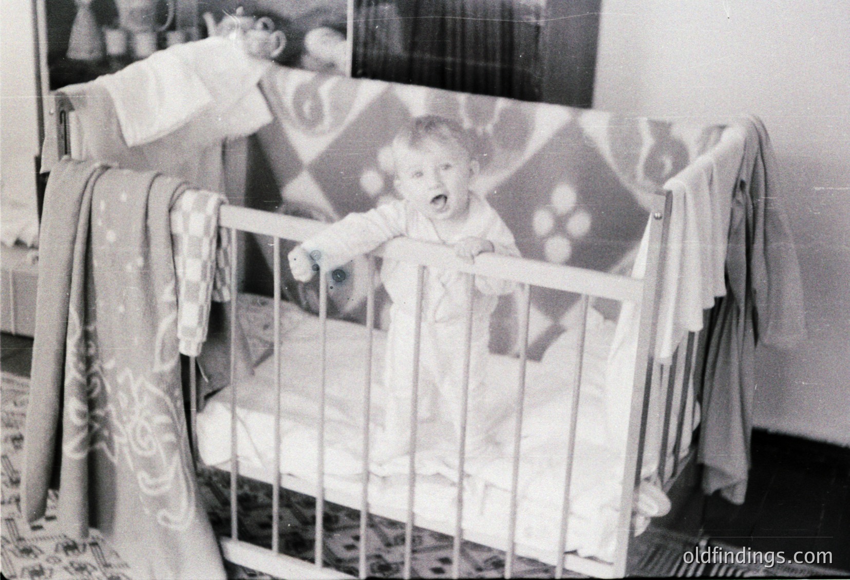 Mid-20th century black-and-white photo of a toddler in a wooden crib with vertical bars, clutching a toy. Patterned wallpaper with floral motifs and a striped blanket in the background. Reflects mid-century domestic life and early childhood play.