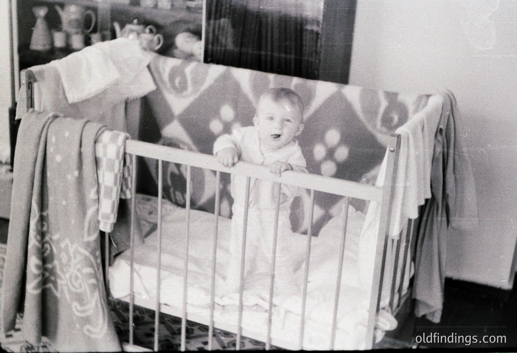 Mid-20th century black-and-white photo of a child in a classic wooden crib with floral-patterned curtains. Baby wears a simple short-sleeve shirt, seated upright. Interior suggests modest, family-oriented home decor. Reflective of mid-century parenting and interior design trends.