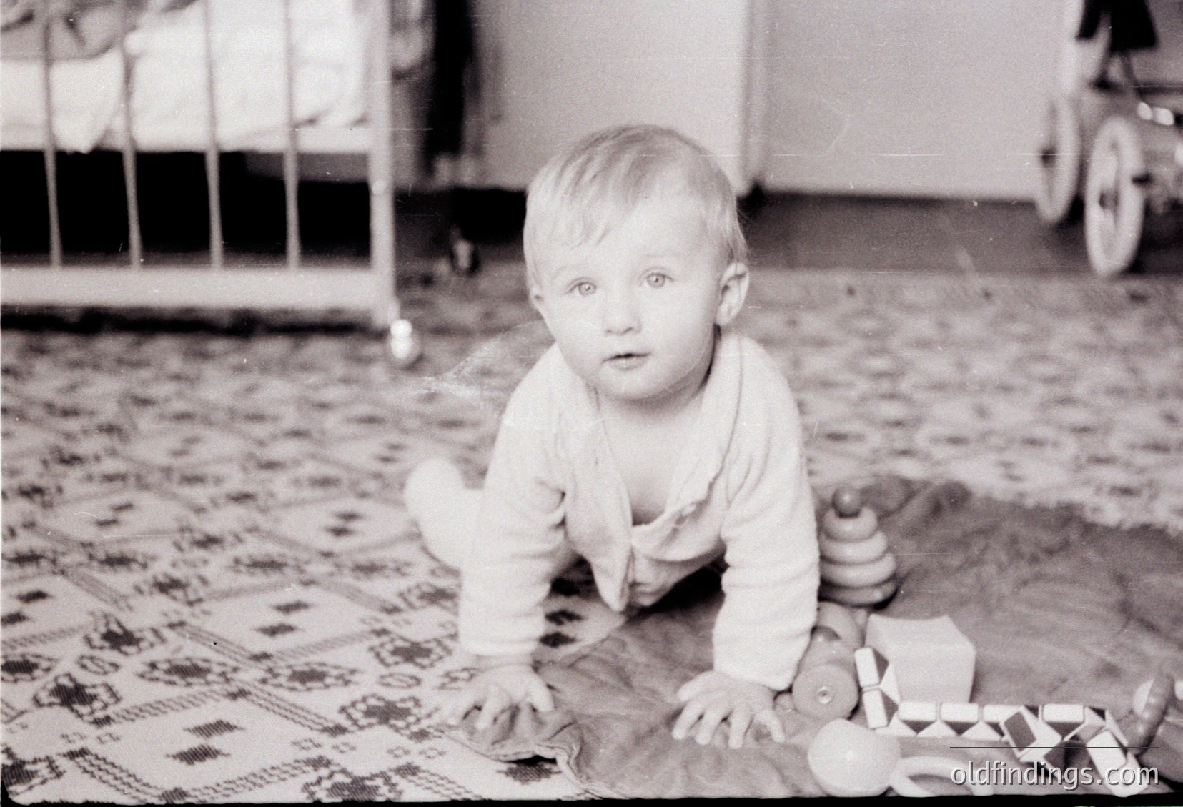 Vintage black-and-white photo of a toddler crawling on patterned rug, surrounded by wooden blocks and a toy train. Mid-20th century domestic setting, likely 1950s–1960s.