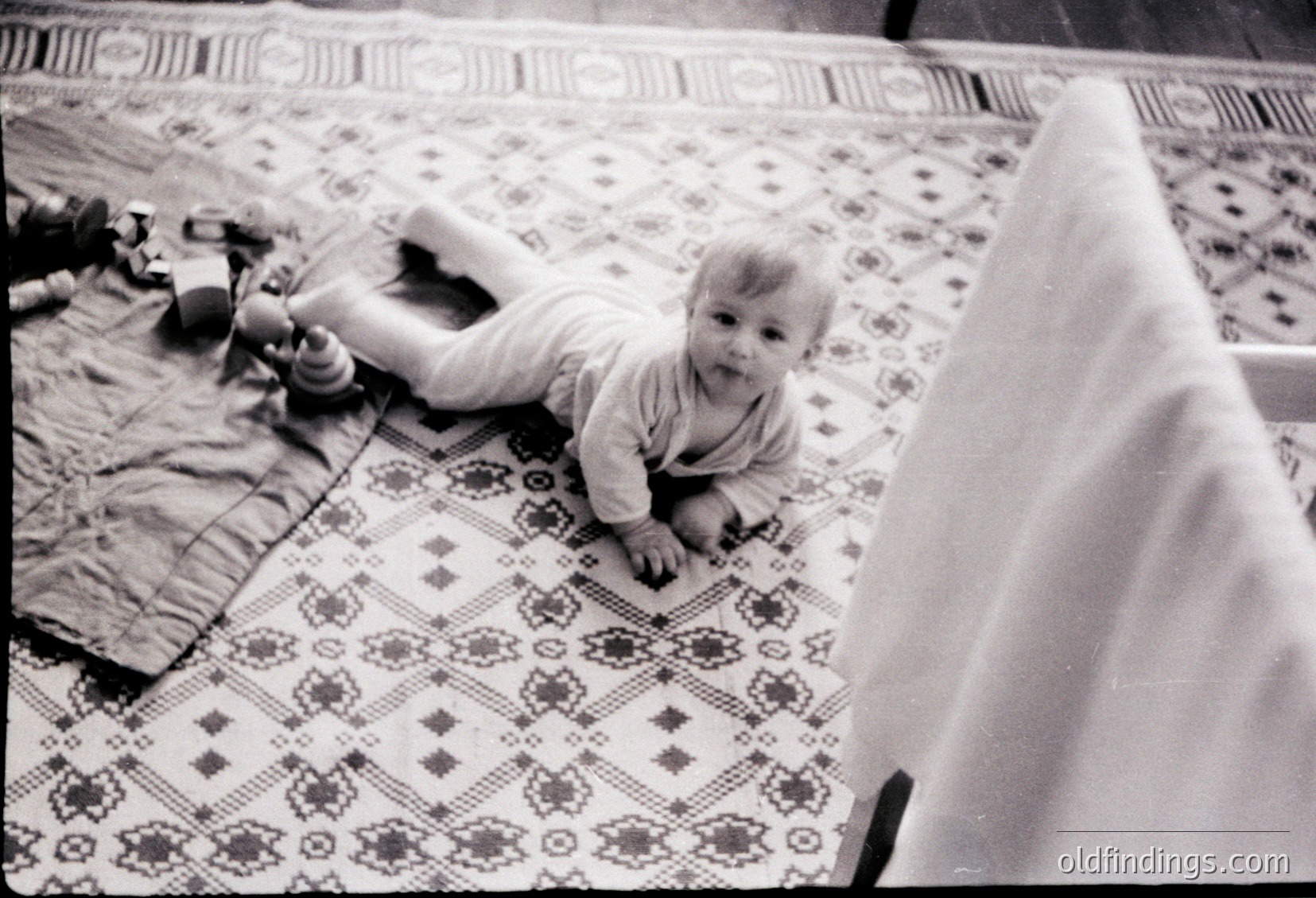 Black-and-white snapshot of a toddler crawling on a patterned rug in a mid-century home. Distinctive floral motifs and geometric designs on the rug suggest 1960s-1970s decor. Baby wears a short-sleeved shirt, playing with a toy near a partially visible crib. Warm, candid family moment.
