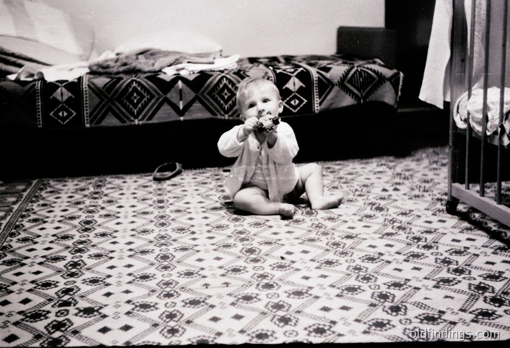 Black-and-white snapshot of an infant crawling on patterned rug, holding a pacifier. Geometric textile designs on bedding and floor suggest mid-20th century domestic style. Warm, intimate family moment captured in vintage aesthetic.