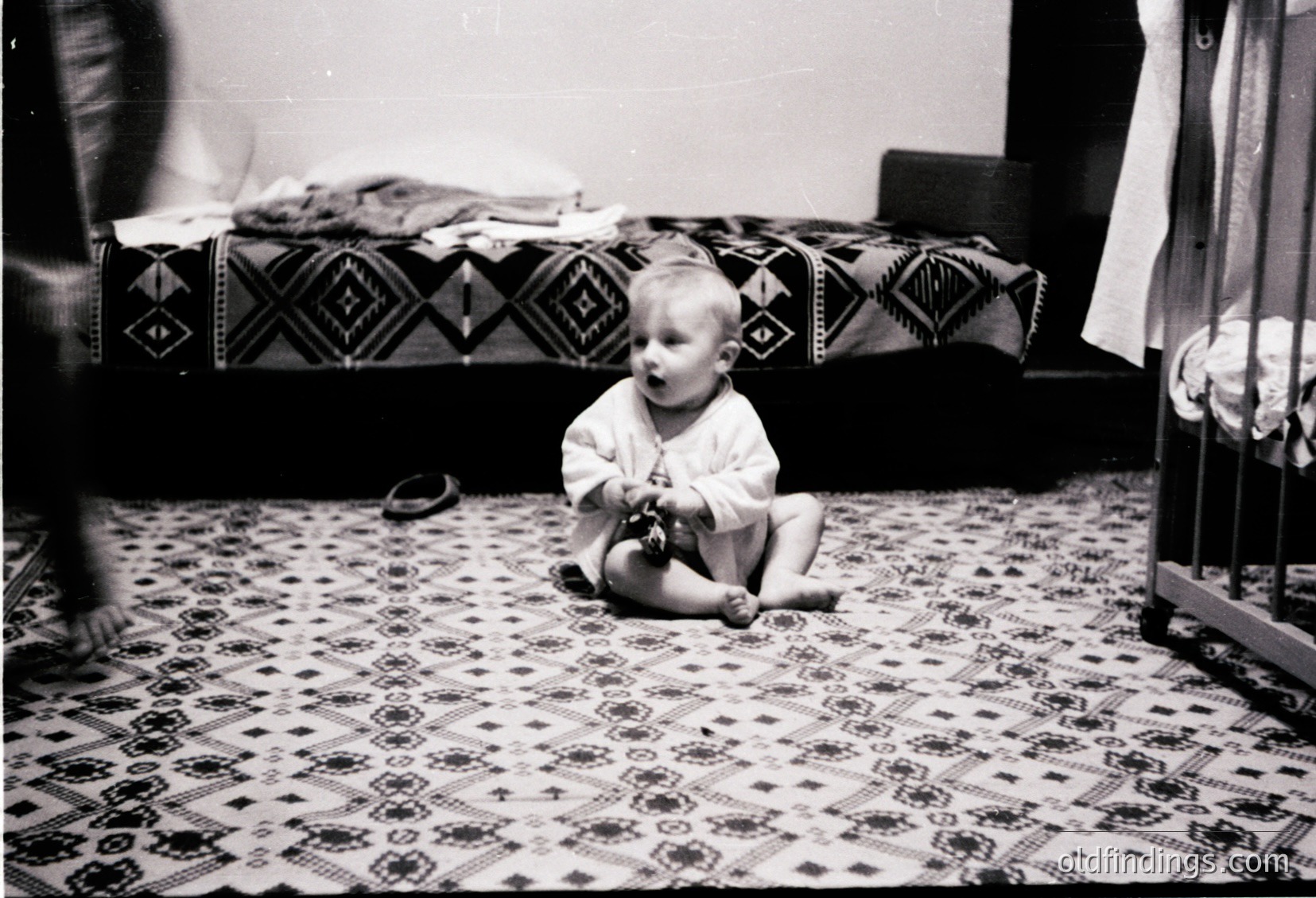 Mid-20th century black-and-white photo of a toddler sitting on patterned rug, playing with a toy. Geometric bedspread with diamond motifs in background. Simple, modest interior suggests domestic setting, likely 1950s–1960s.