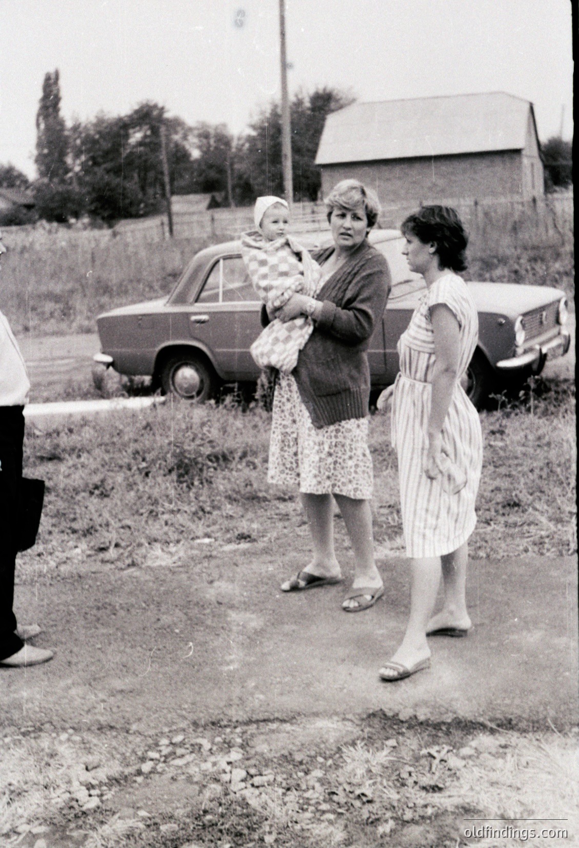 Mid-20th century rural scene featuring three women: one holding a baby in a floral dress, another in a striped dress carrying groceries, and a third in a white blouse. Classic vintage car (likely a Lada or similar) parked on unpaved road. Rustic farmhouse and trees in background.