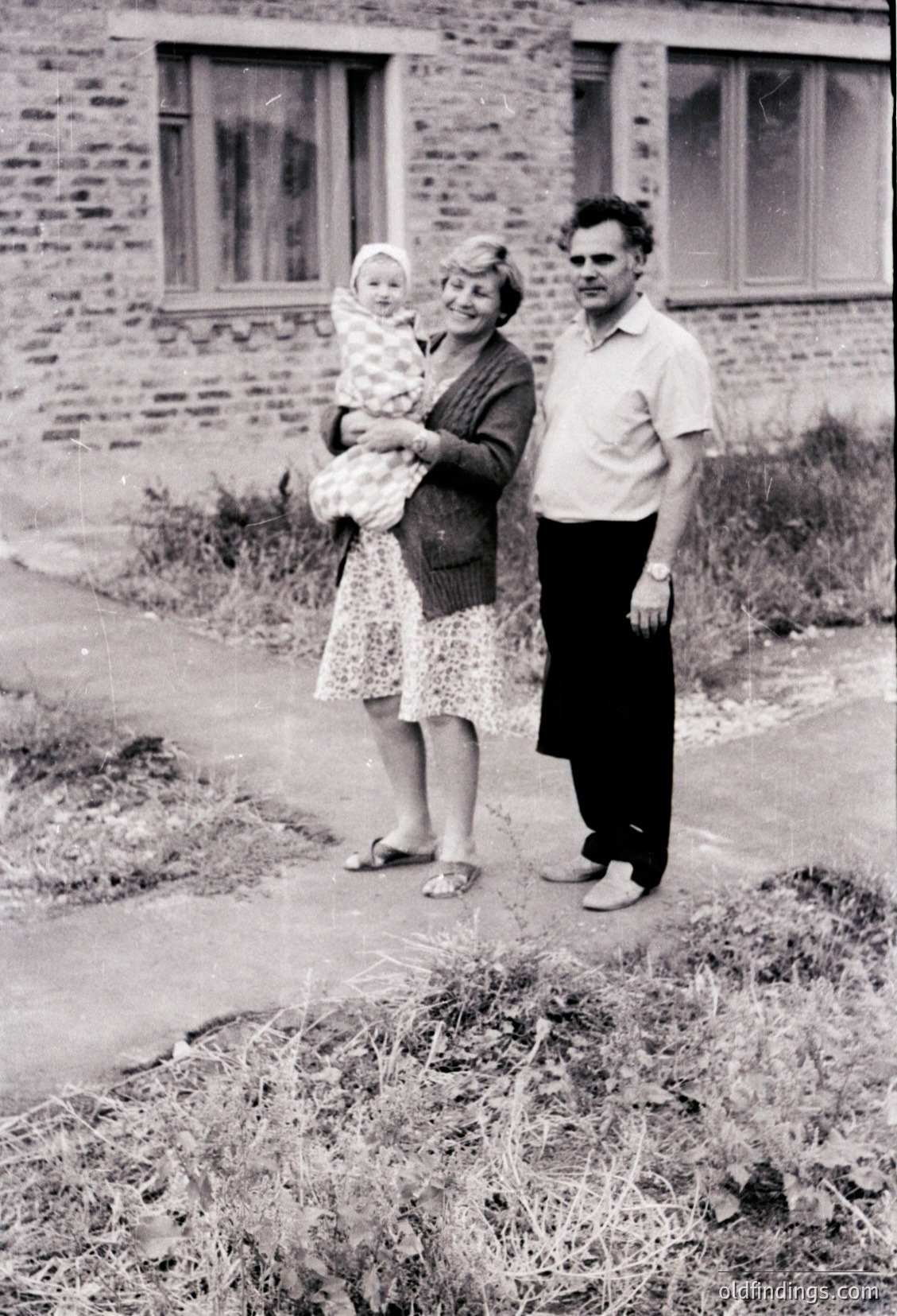 Family portrait in front of a brick residential building, likely Eastern Bloc-era (1960s–1970s). Woman holds a baby in a patterned dress, flanked by a man in a short-sleeve shirt. Overgrown grass and unpaved path suggest rural or suburban setting.