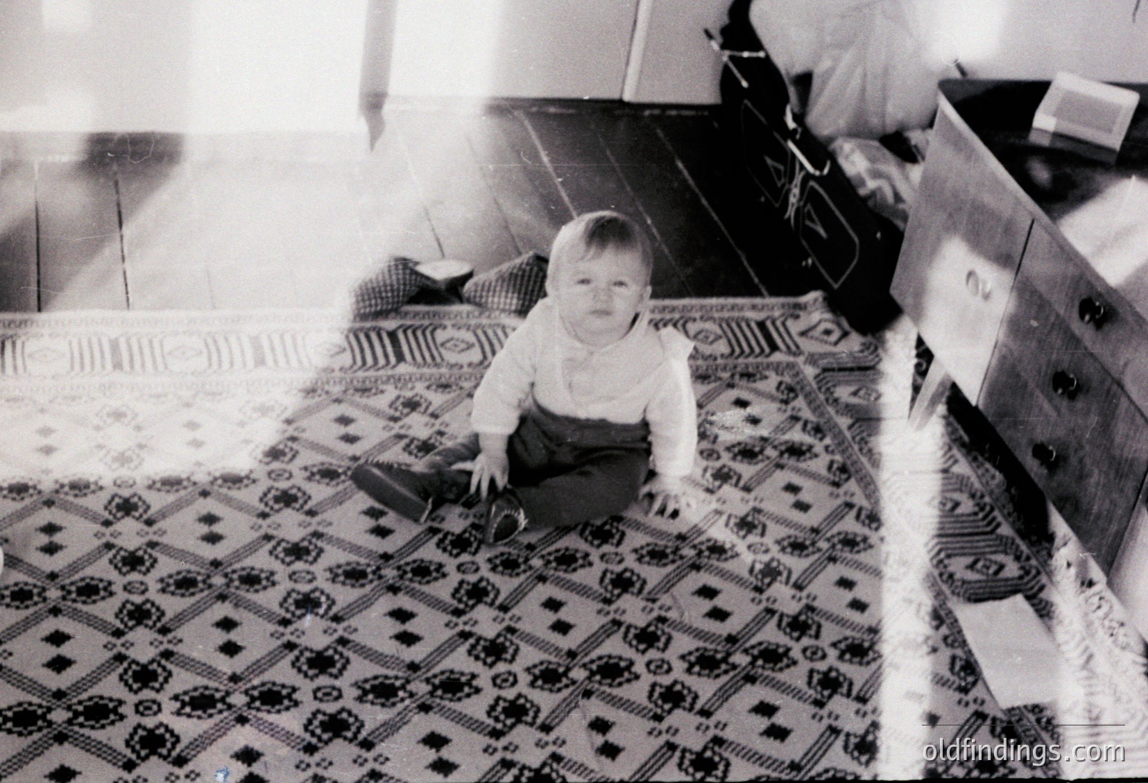 A toddler sits cross-legged on a patterned rug with geometric and floral motifs, wearing a short-sleeved shirt and pants. The vintage black-and-white photo captures a mid-20th-century domestic interior, likely or , with a wooden cabinet and partial view of a chair. The child’s expression and setting evoke nostalgia for family life.