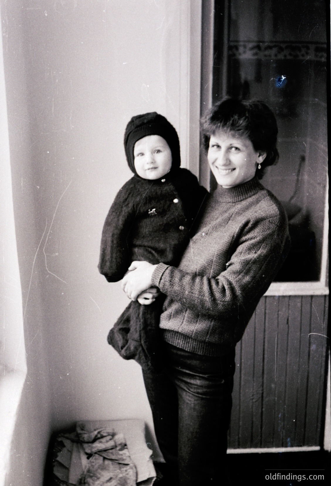 Black-and-white portrait of a woman cradling a toddler indoors, likely mid-20th century. The woman wears a knitted sweater with a buttoned collar and dark pants; the child is bundled in a knit hat and jacket. Plain wall and window frame suggest a modest residential setting.