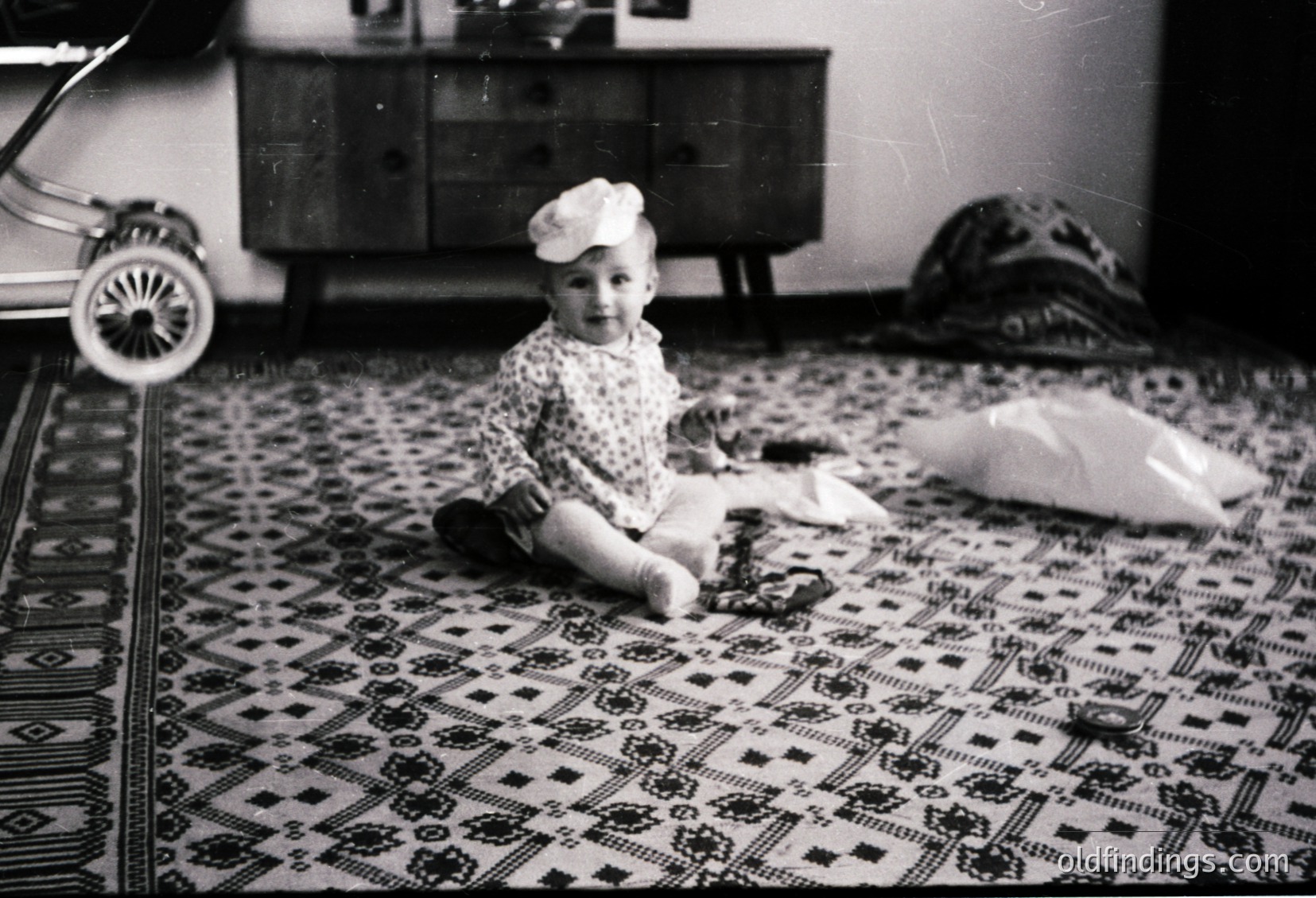 Vintage black-and-white photo of a toddler sitting on an intricately patterned rug in a mid-century home. The child wears a polka-dot dress and a white hat with a brim. Mid-century furniture, including a wooden dresser and a toy stroller, fills the background. Likely 1950s–1960s domestic setting.