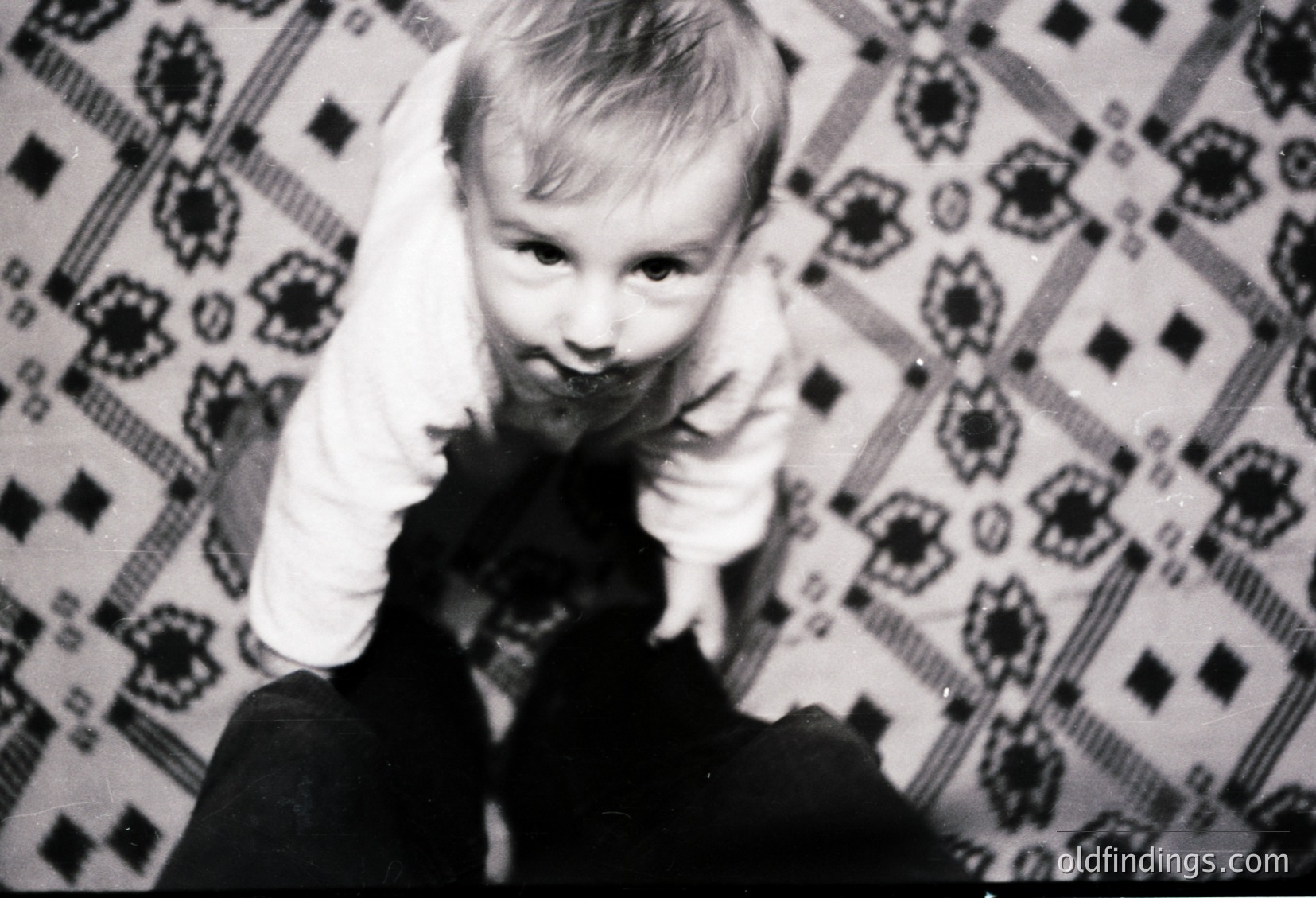 Vintage black-and-white photo of a child making a peace sign against a patterned wallpaper backdrop. Mid-20th century indoor setting, likely residential. Candid, playful expression captures youthful energy.