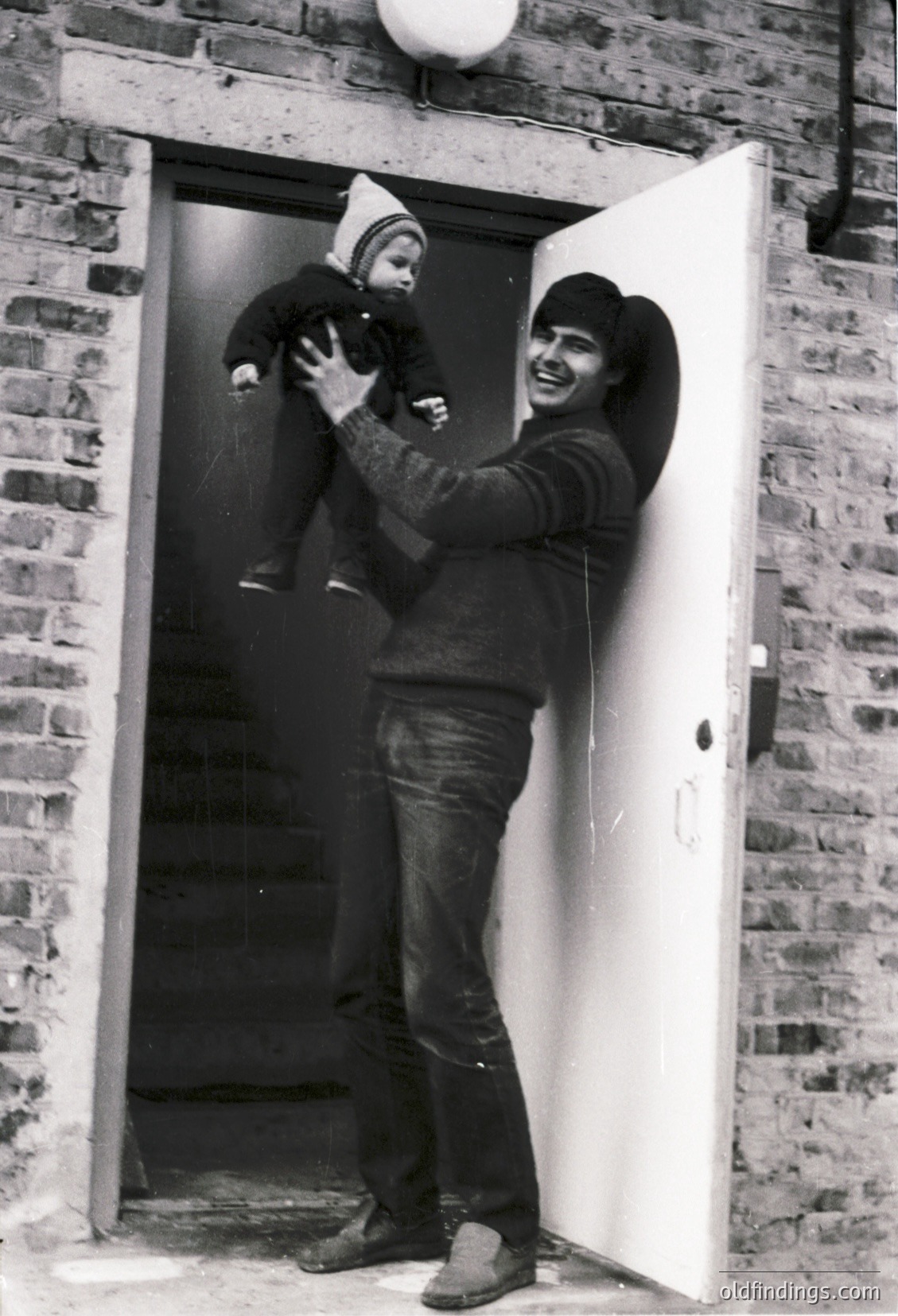 Mid-20th century black-and-white photo of a man holding a toddler at a brick-framed doorway. The man wears a cap, sweater, and jeans; the child wears a knitted hat and coat. Warm, candid family moment. Likely