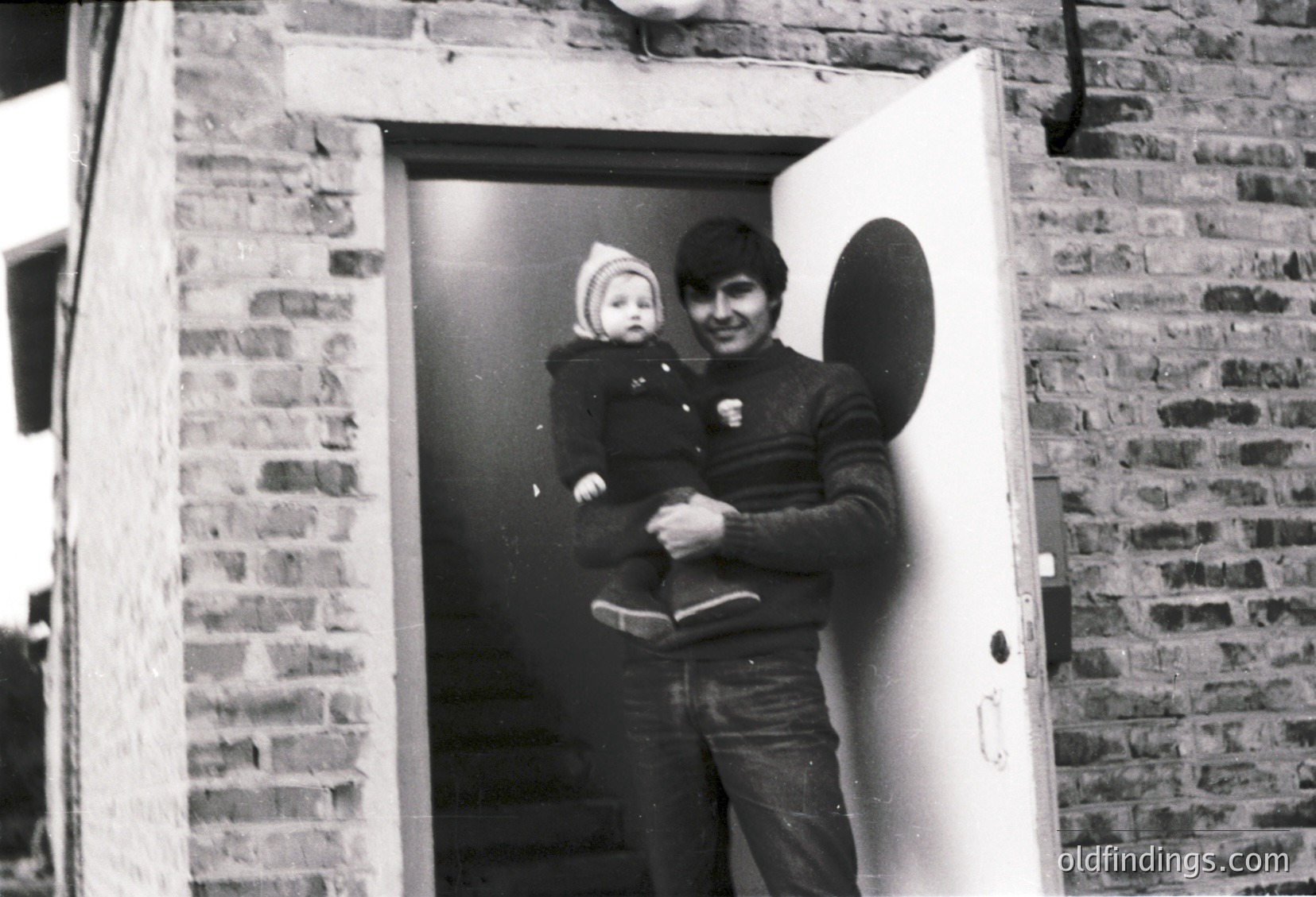 A man in mid-20th-century attire holds a baby at a brick entranceway, framed by a white door. The man wears a dark jacket with a patch, jeans, and a belt; the baby is bundled in a knitted hat and coat. The brickwork suggests an urban residential setting, likely –. Ideal for nostalgic or family history research.