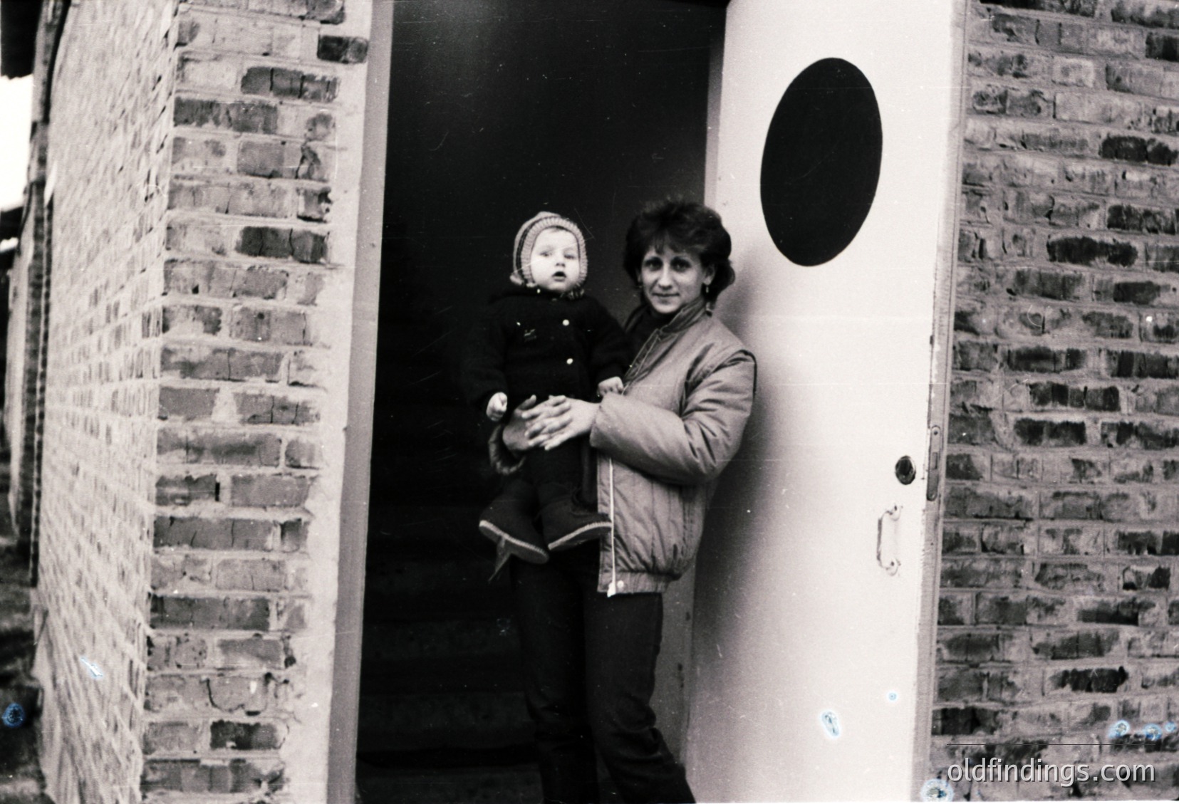 A woman holds a toddler in a brick-lined hallway, framed by a white door with a black circular design. Mid-20th century urban setting, likely Eastern Europe.