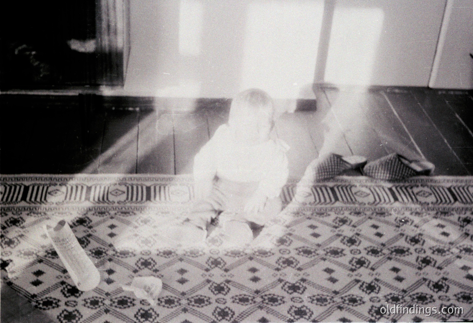Vintage black-and-white photo of an infant playing on a patterned rug with geometric floral motifs. Soft focus reveals a minimalist interior with tiled floor and a partially visible door. Likely mid-20th century domestic setting.