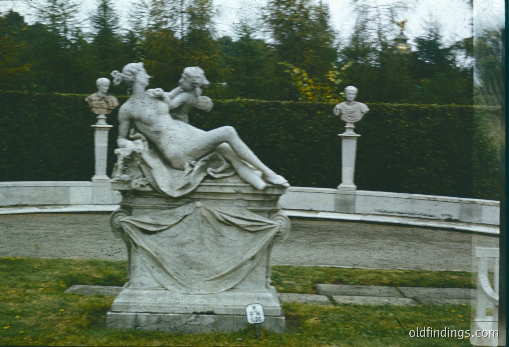 Neoclassical stone fountain sculpture depicting reclining female figure with draped drapery, flanked by two busts on pedestals. Set in a formal garden with symmetrical stone steps and lush greenery. Likely European, 18th–19th century.