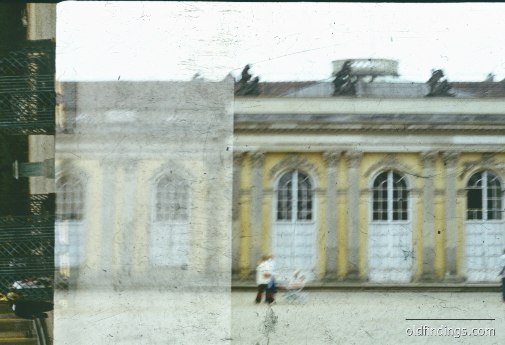 Vintage sepia-toned photo of a grand neoclassical building with arched windows and cream/yellow façade, likely mid-20th century. A lone figure in light clothing walks near the entrance, framed by a slightly blurred foreground. Architectural details suggest institutional or governmental use.
