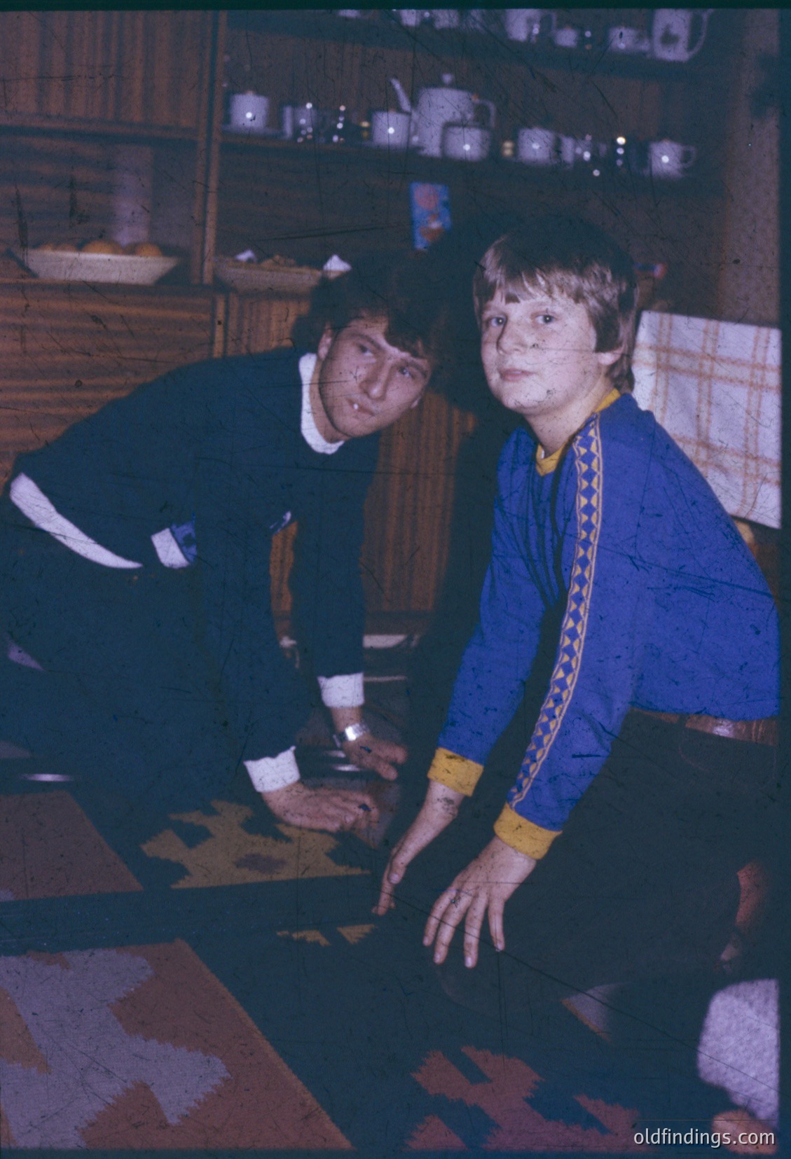 Two individuals in 1970s-style sportswear—blue and yellow tracksuits—lean over a patterned carpet, examining a small object on a wooden table. Shelves behind them hold ceramic mugs and vintage kitchenware. Warm, indoor lighting suggests a domestic or training setting.