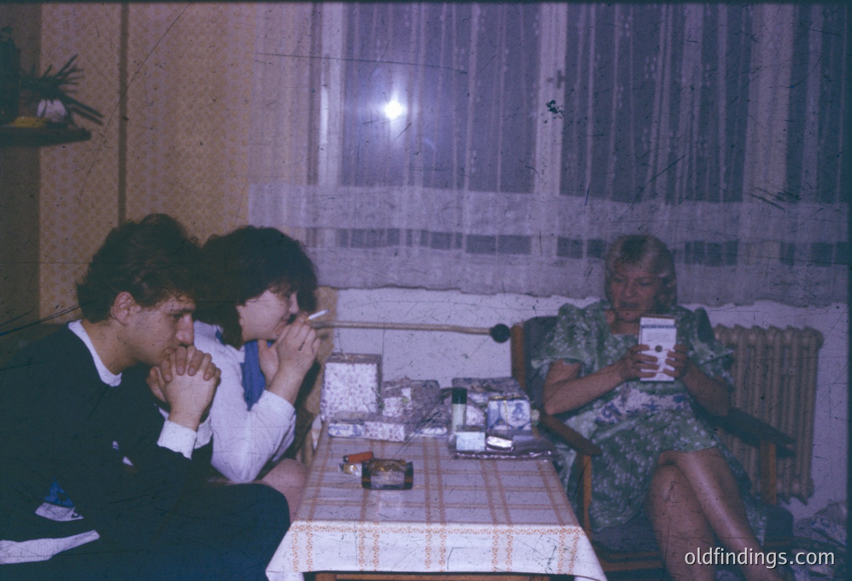 Three individuals seated indoors in a dimly lit room, likely mid-1970s–1980s Eastern Bloc. Man in suit (left) holds a badge, woman (center) wears a blue scarf, woman (right) in floral dress drinks. Tabletop features framed photos, a radio, and a small plant. Heavy curtains and radiator suggest utilitarian Soviet-era apartment decor.